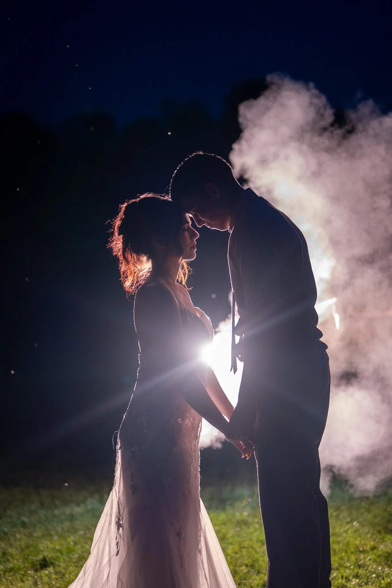 Silhouette of a couple embracing outdoors at night, with steam or smoke in the background, backlit by bright light creating a romantic glow.