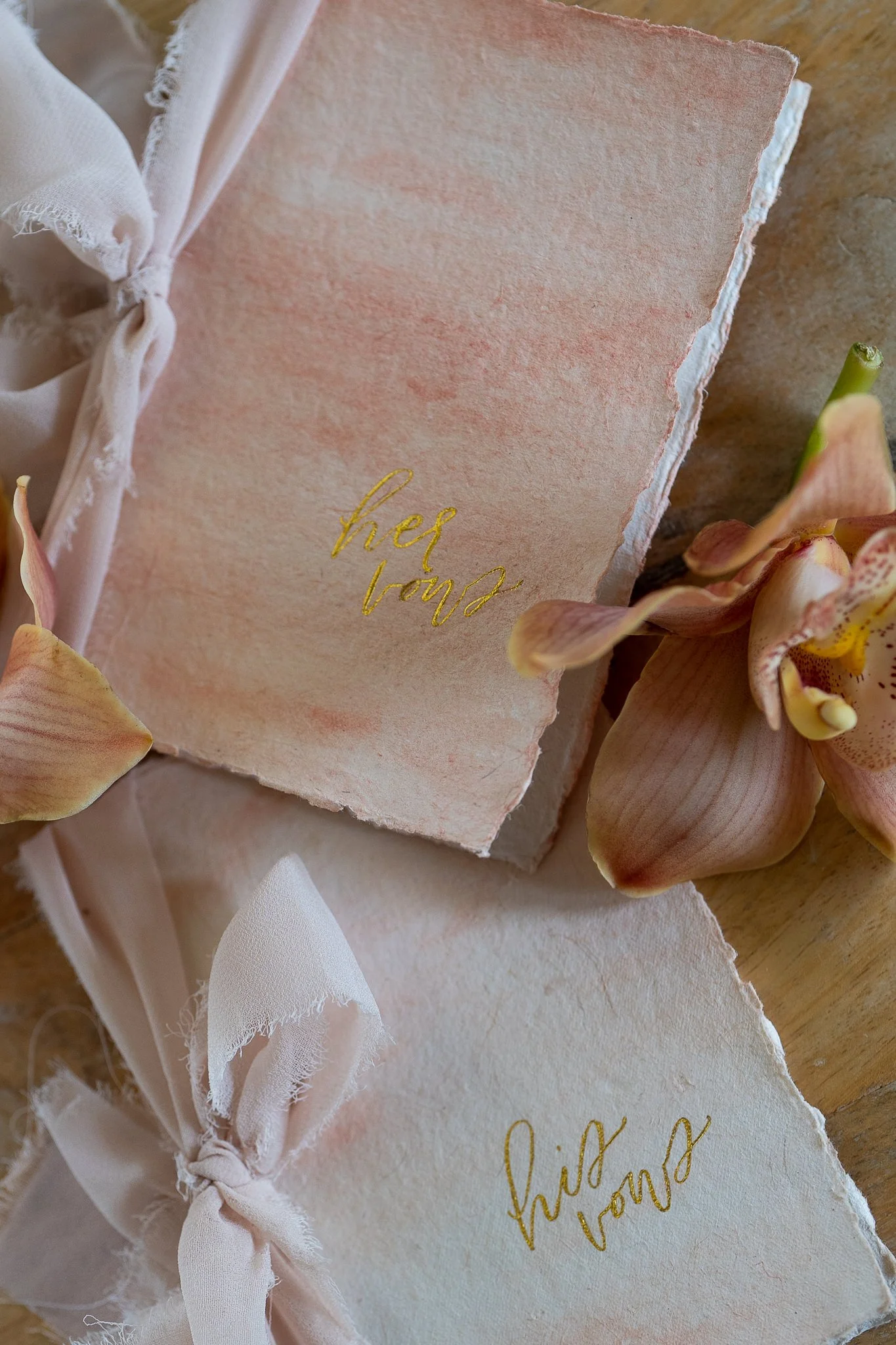 Wedding invitation cards with gold script, surrounded by pink orchids and light-colored fabric tied with bows.