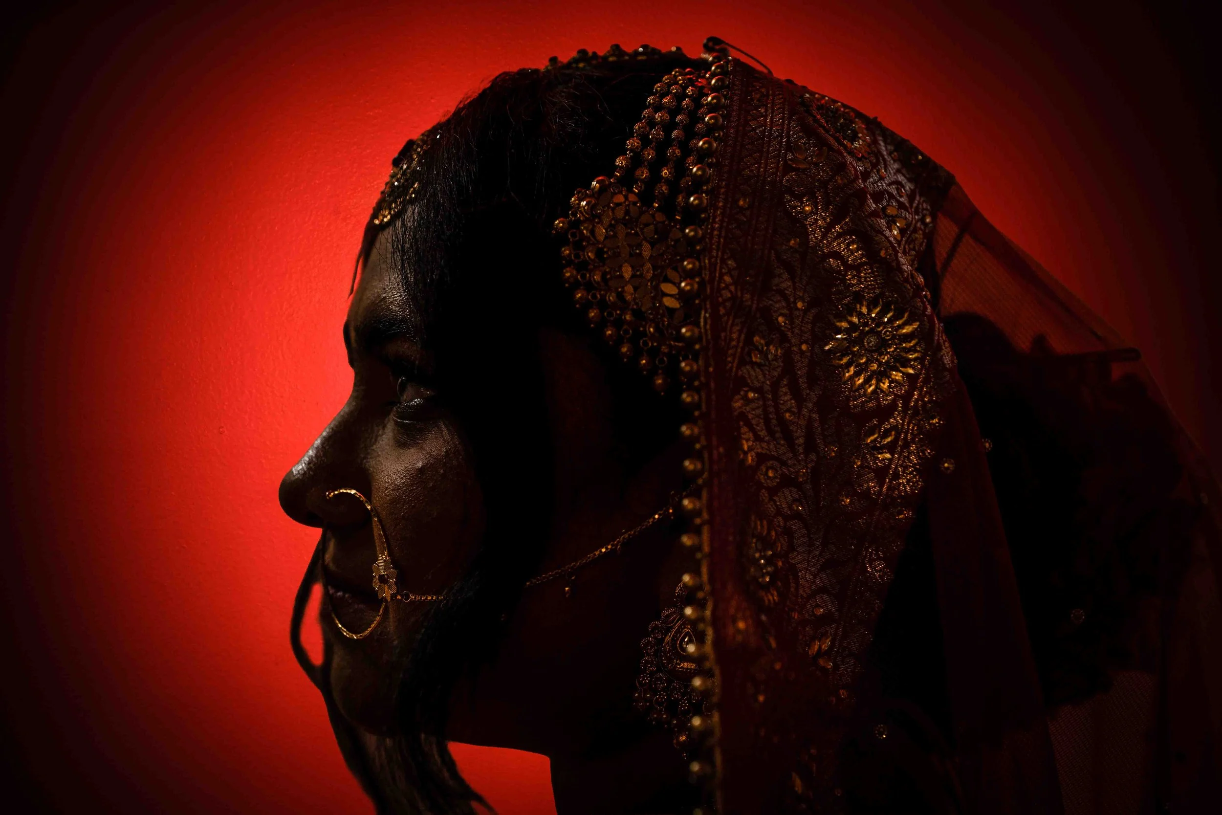 A profile of a woman wearing traditional jewelry and a richly embroidered veil, illuminated by warm red lighting.