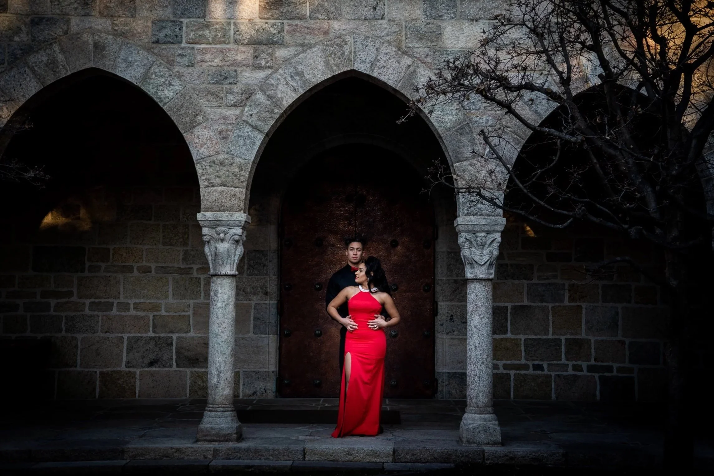 A man and woman are standing together in front of a large wooden door framed by stone arches. The woman is wearing a red evening gown with a slit, and the man is in dark clothing. The setting appears to be outside an old stone building, with leafless