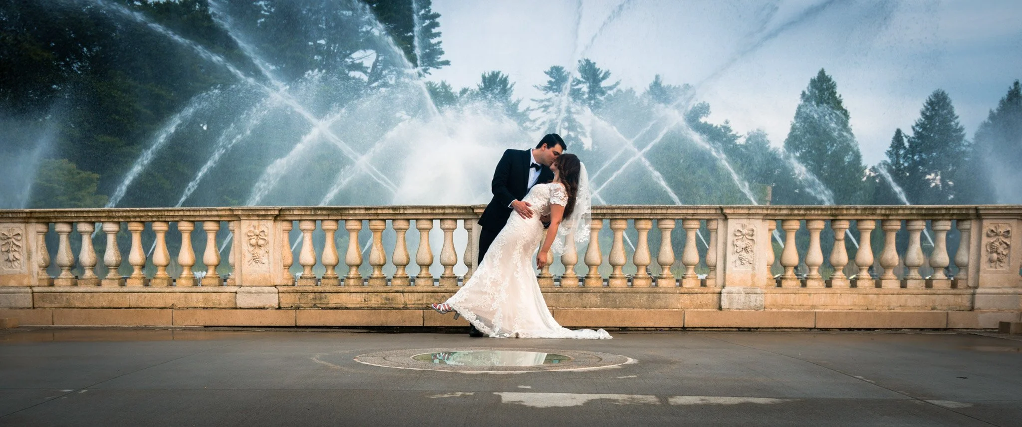 A couple dressed in wedding attire sharing a kiss in front of a stone railing with a large fountain and trees in the background.