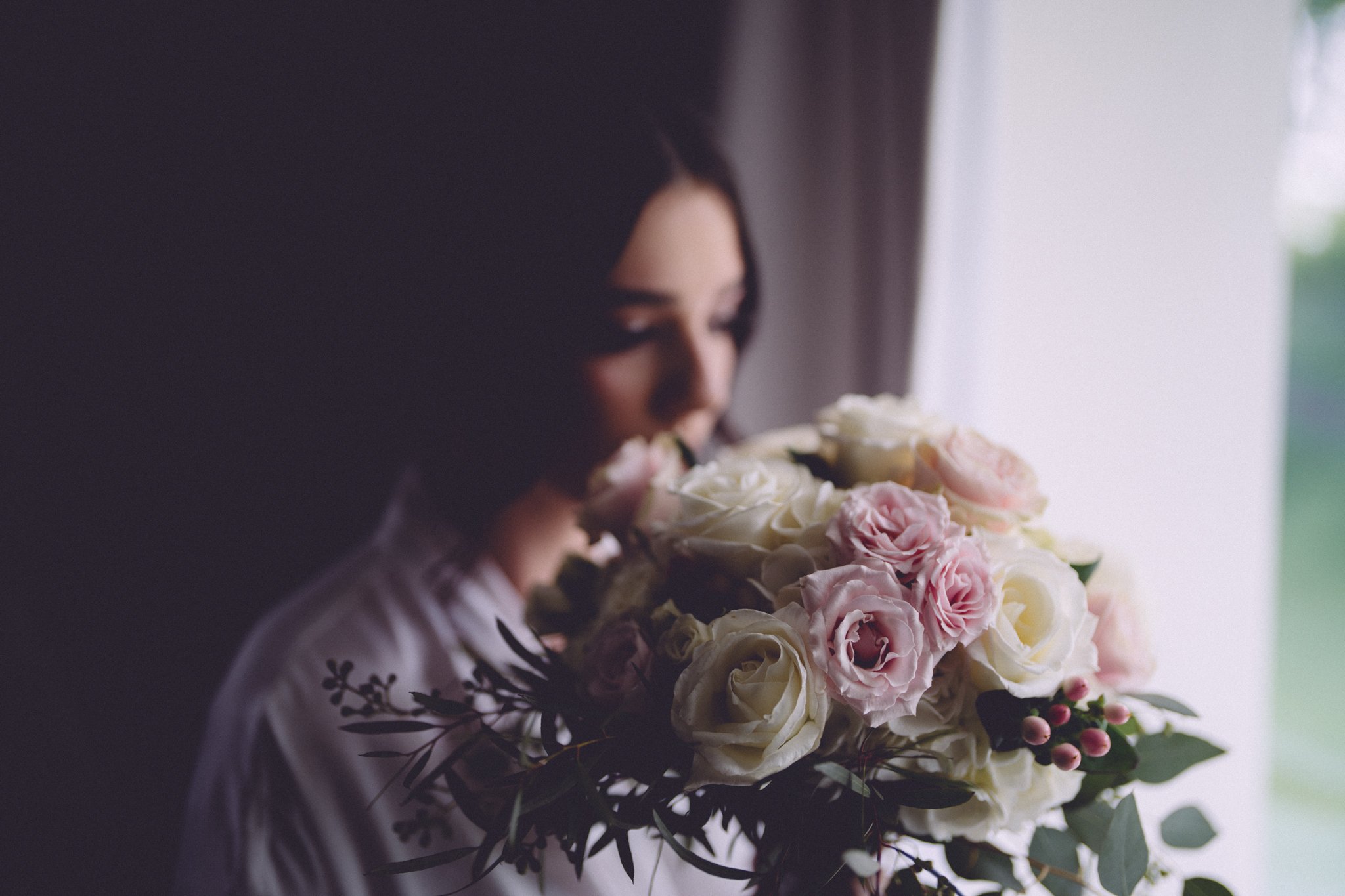 A woman with black hair holding a large bouquet of pale pink and white roses near her face, standing near a window with light shining in.