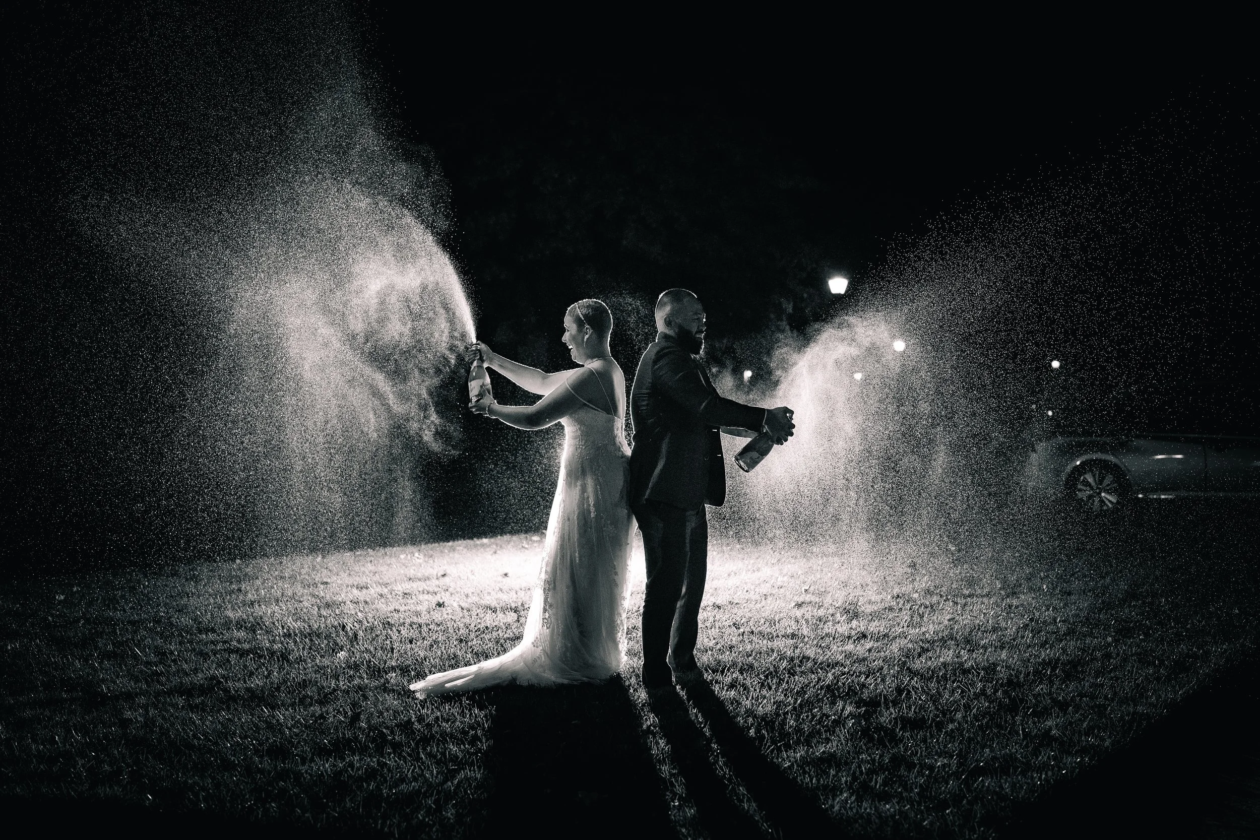 A black and white photo of a bride and groom celebrating at night, with the bride spraying champagne and the groom holding a champagne bottle, surrounded by lights and parked cars.