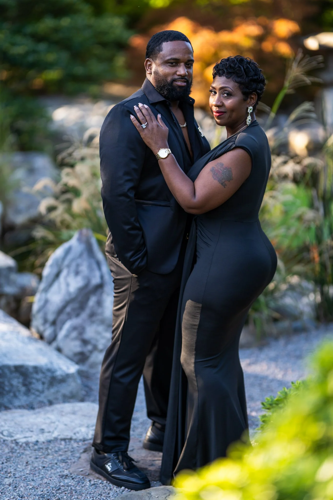 A man and woman dressed in black, standing close together outdoors during sunset, embracing and posing for a photo, with trees and rocks in the background.