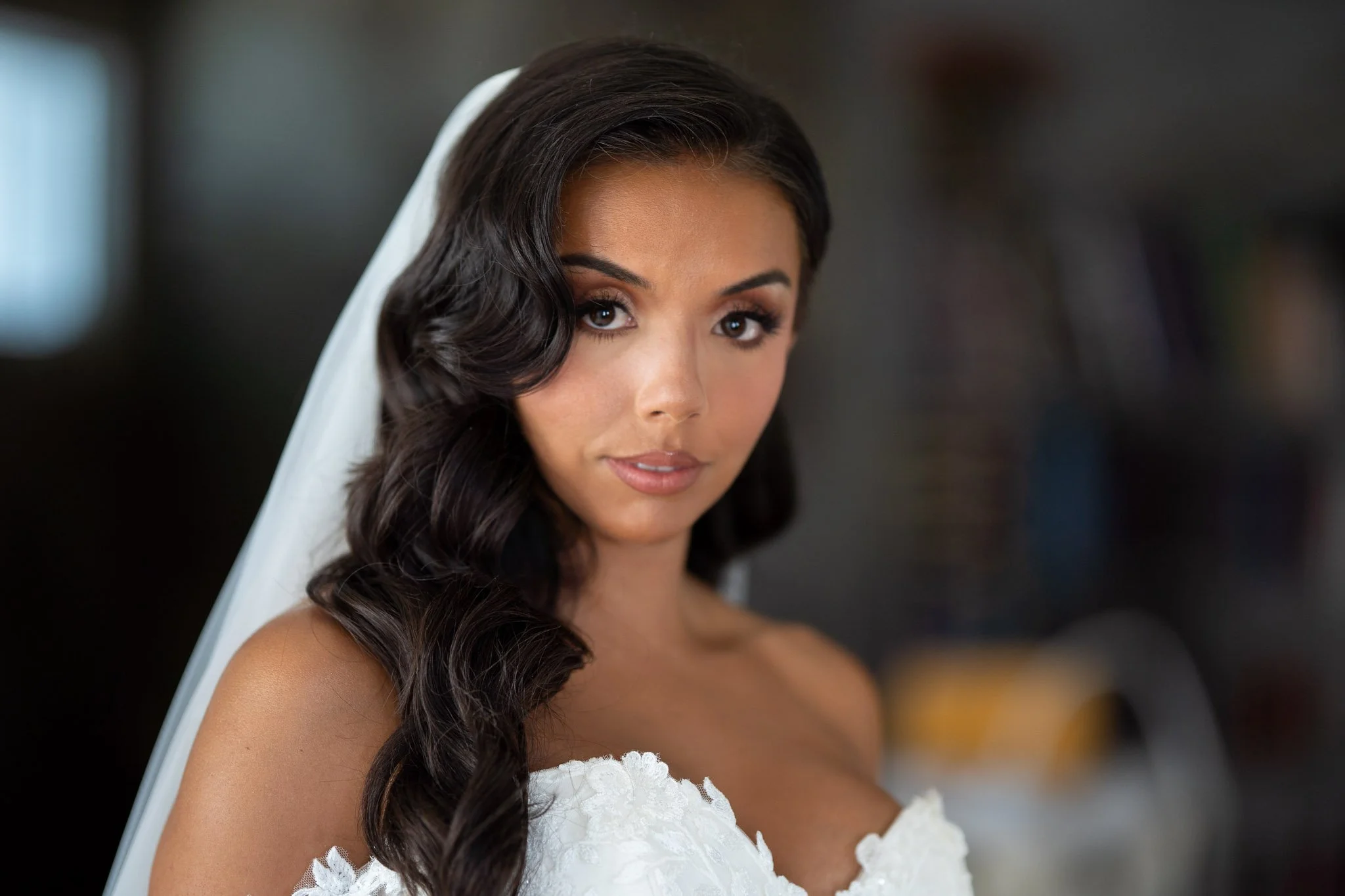 Close-up of a young woman with dark, wavy hair, and makeup, wearing a white dress with lace details on the shoulder, and a white veil, looking at the camera.