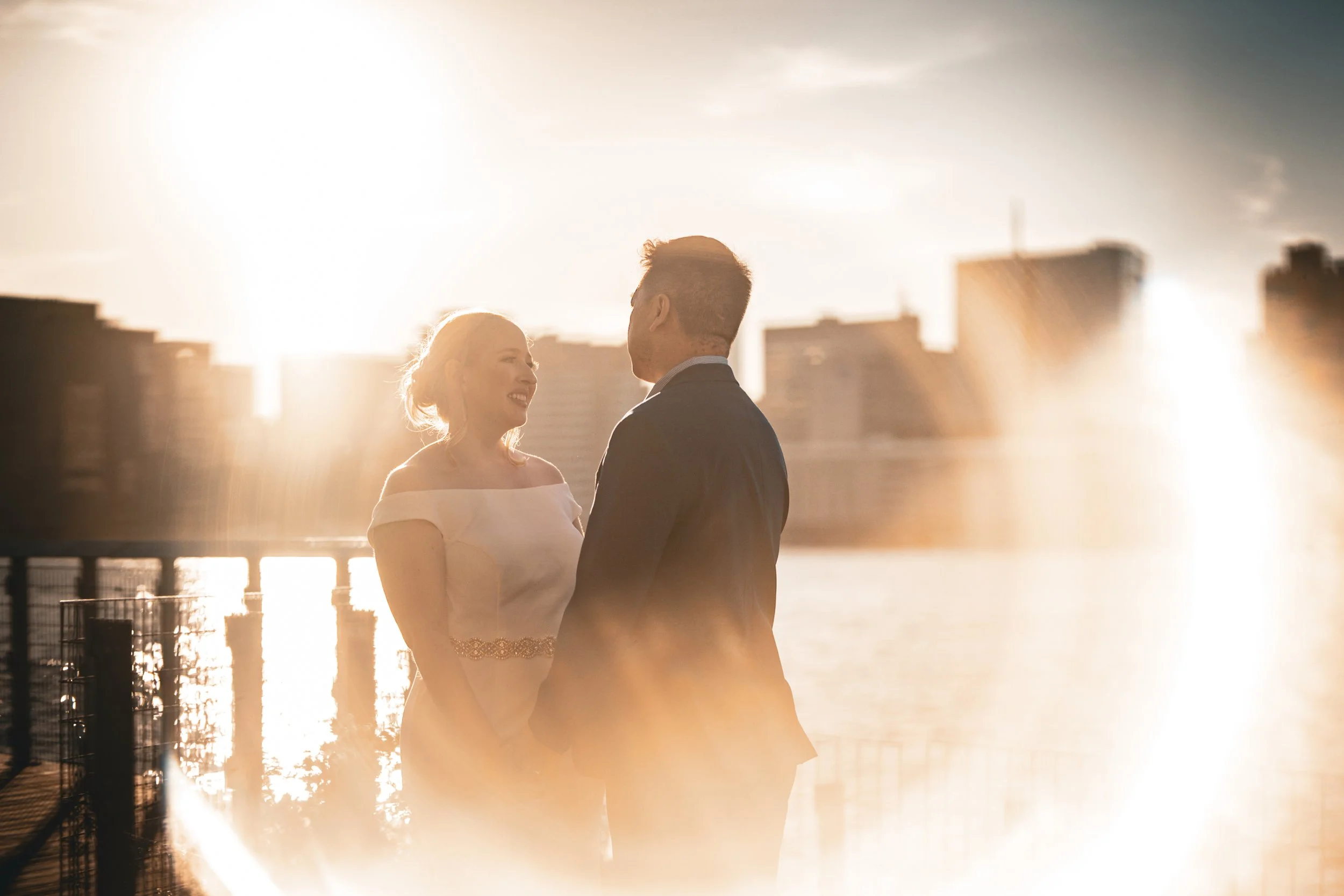 A couple in wedding attire holding hands and looking at each other with city skyline and river in the background during sunset.