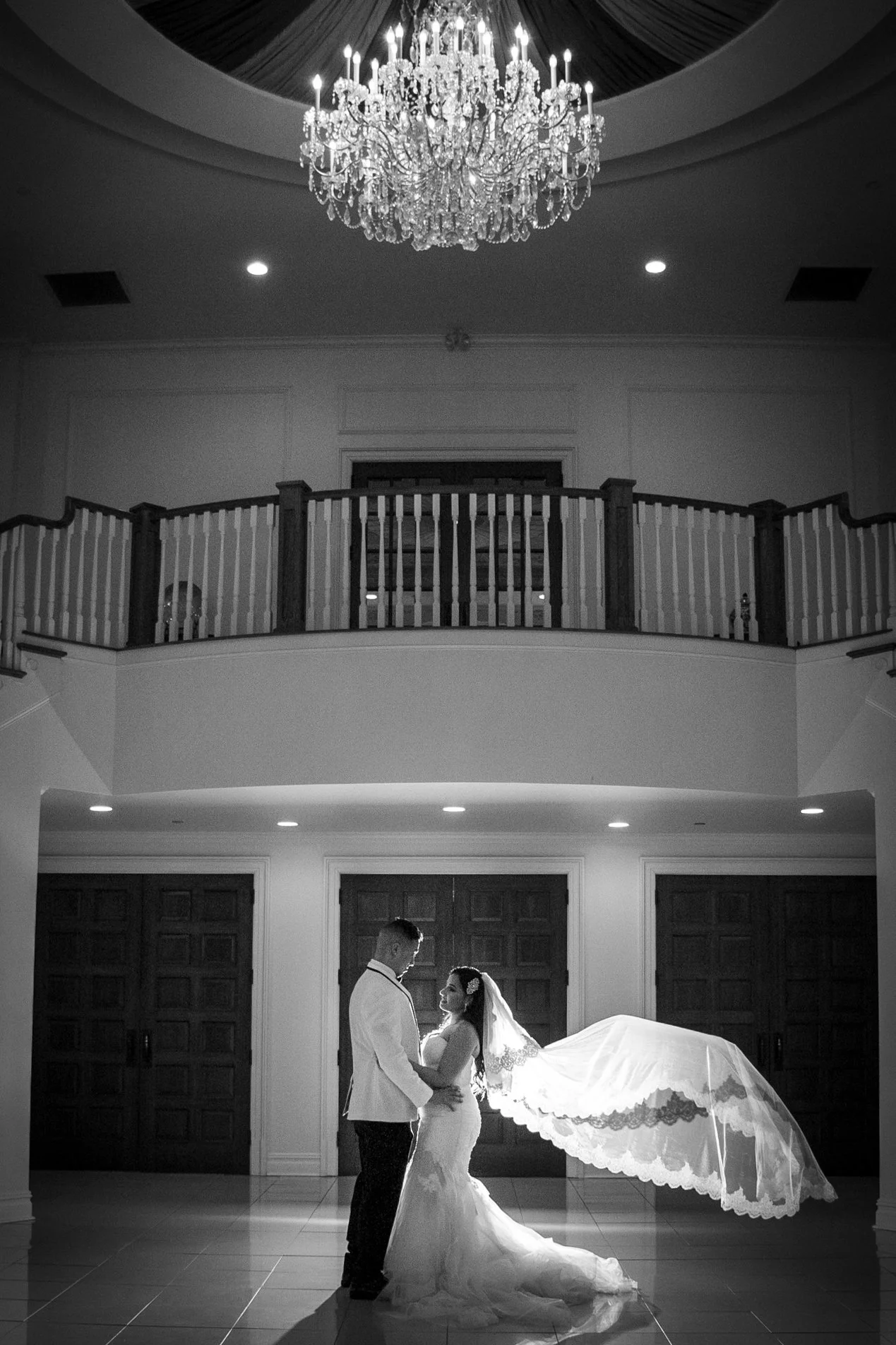 A bride and groom standing close together, holding hands, in a grand indoor setting with a chandelier overhead and wooden double doors in the background.
