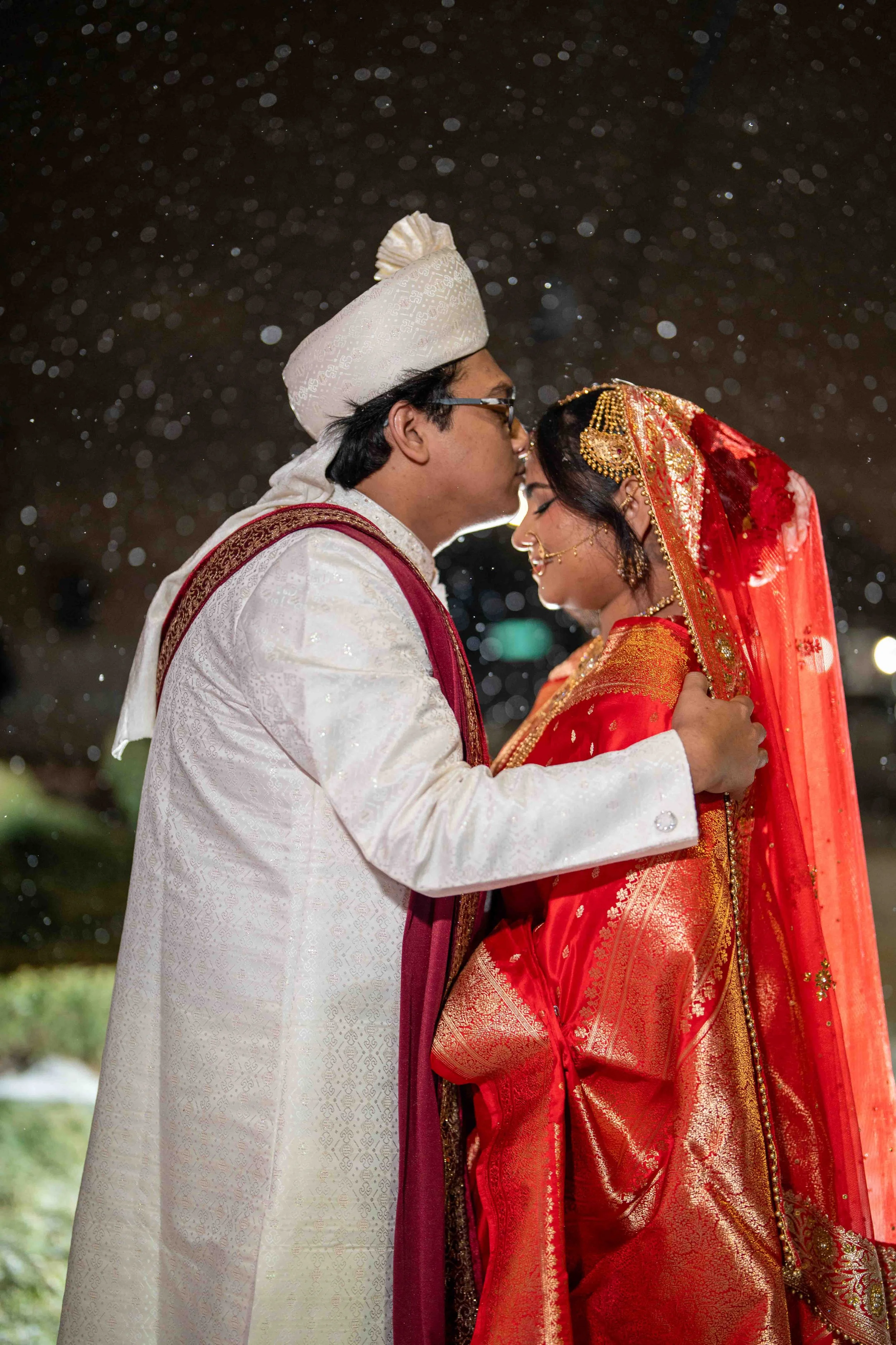 A couple dressed in traditional Indian wedding attire, standing close with foreheads touching, during a rain shower at night.