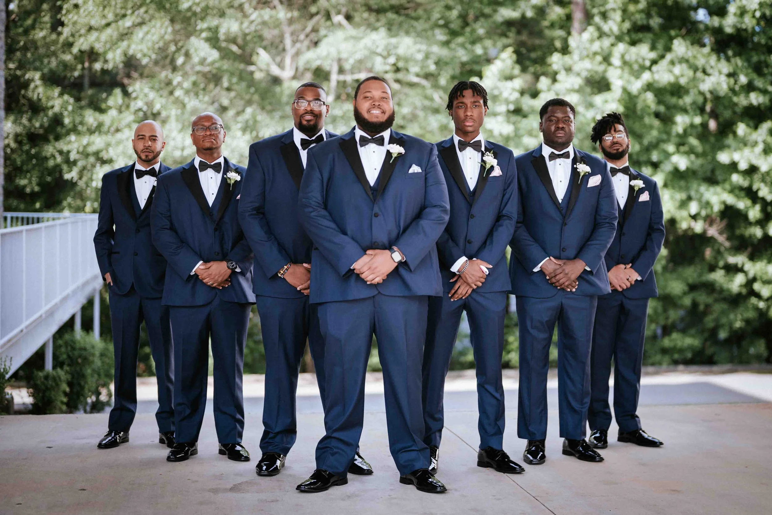 A group of seven men dressed in matching dark blue tuxedos with black lapels, white shirts, black bow ties, and matching shoes, standing outdoors on a concrete pathway with green trees in the background, posing for a formal photo.