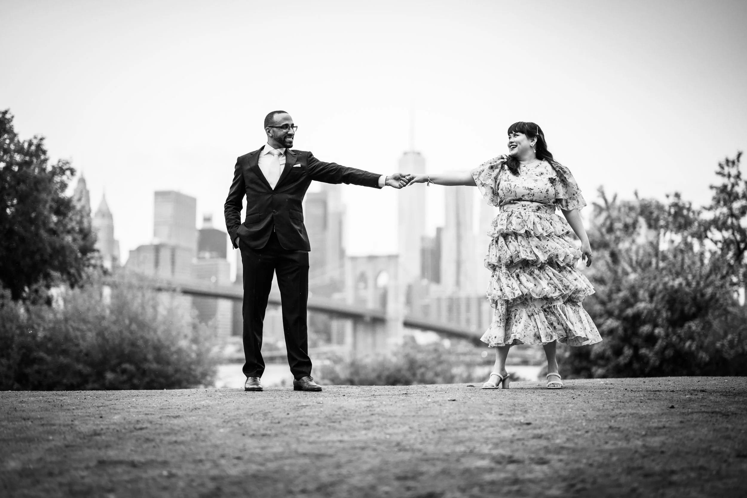 A black-and-white photo of a man and woman holding hands in a city park with Brooklyn Bridge and Manhattan skyline in the background. The man is dressed in a suit, and the woman is wearing a floral dress with ruffles.
