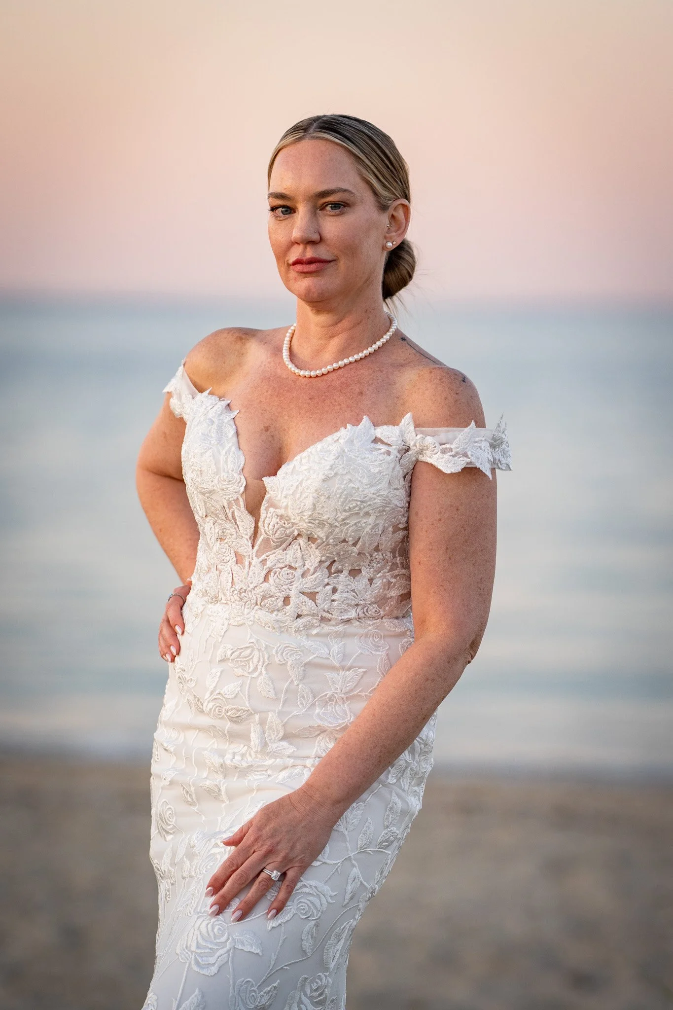 A woman in a white lace wedding dress standing by the beach at sunset.