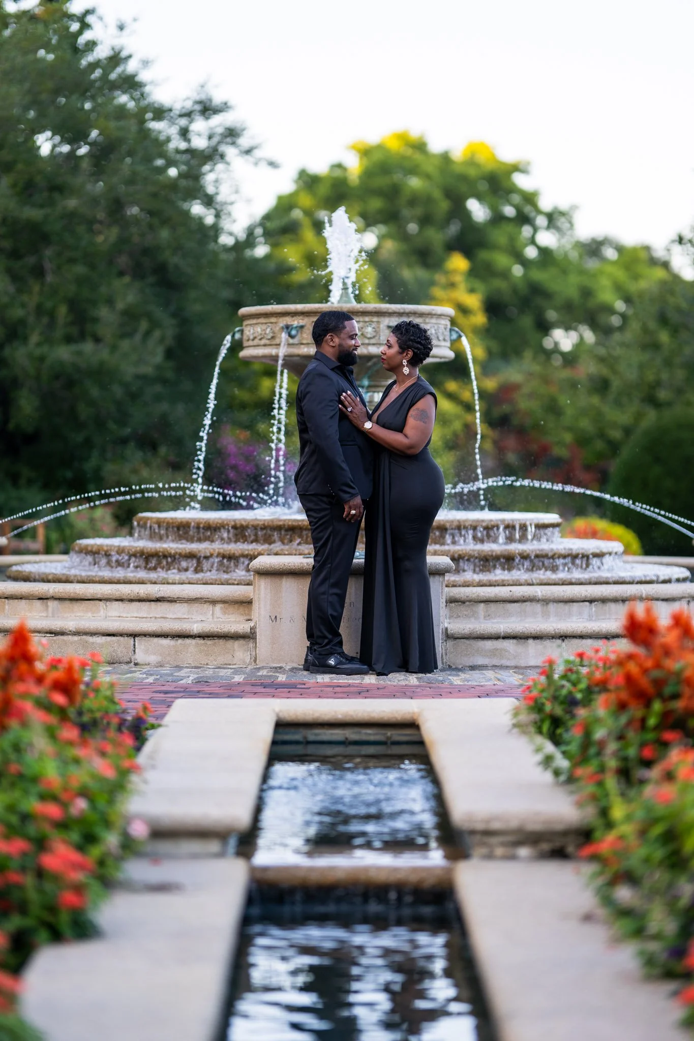 A couple dressed in black stands close together in front of a decorative fountain with water cascading from multiple tiers in a park setting during daylight. The woman has short hair and earrings, and the man has a beard and dark hair. The background