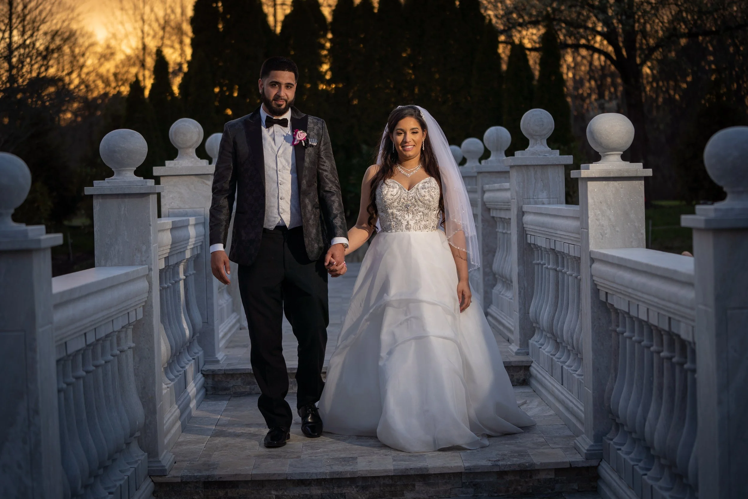 A newlywed couple walking on a marble bridge at sunset. The groom is wearing a black tuxedo with a bow tie and boutonnière, and the bride is in a white wedding gown with intricate embroidery and a veil. They are holding hands and smiling, with trees 