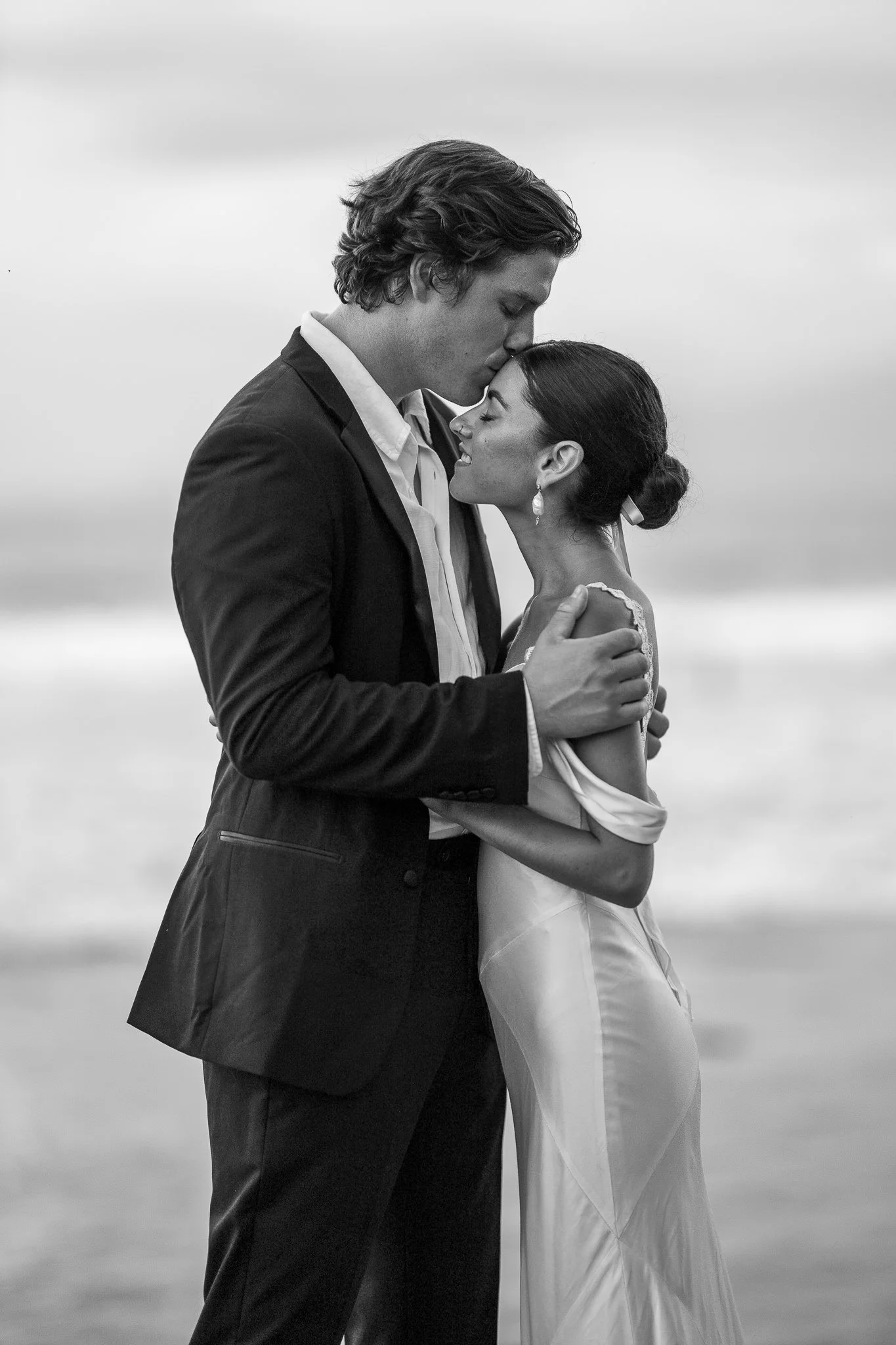 A black-and-white photograph of a man and woman embracing on a beach. The man is kissing the woman's forehead, and they are both dressed formally, the woman in a gown and the man in a suit.