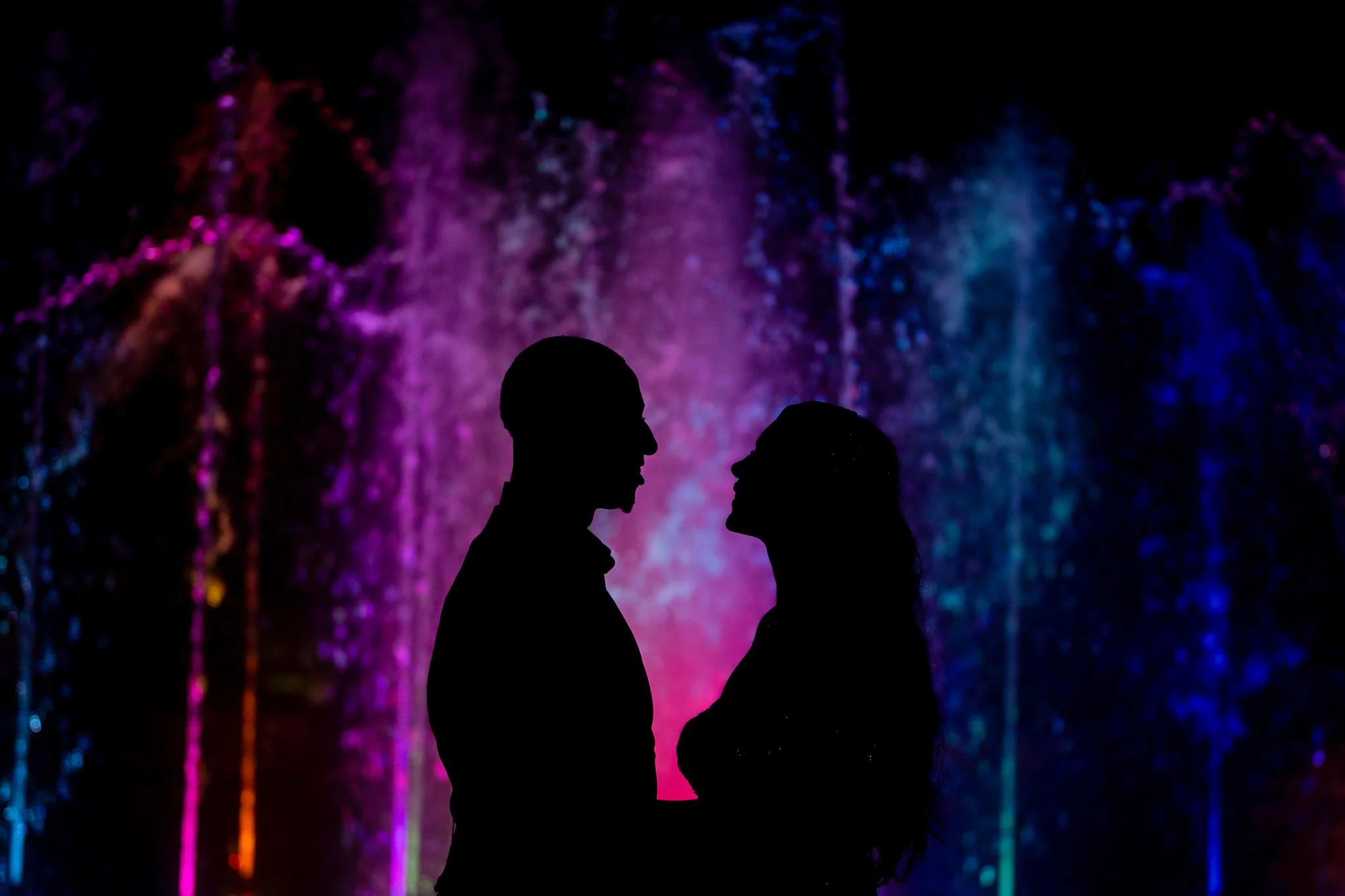 Silhouettes of a man and woman facing each other against a colorful illuminated water fountain background.