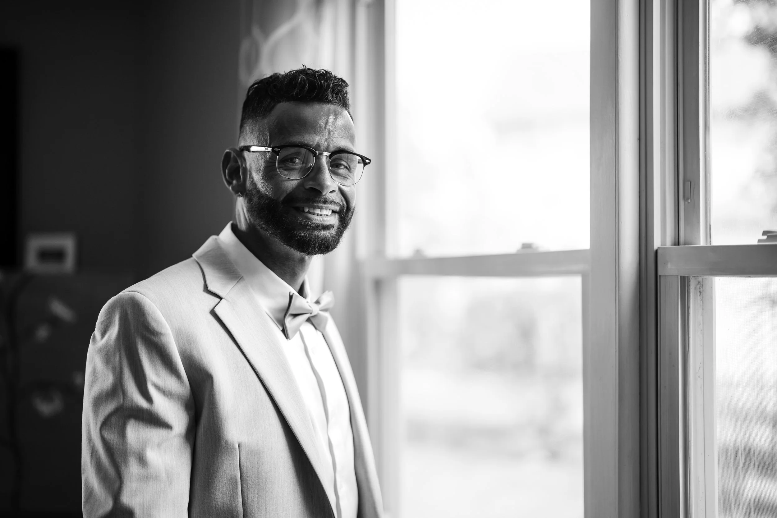 A man in a tuxedo and bow tie standing next to a window, smiling at the camera, in black-and-white photography.