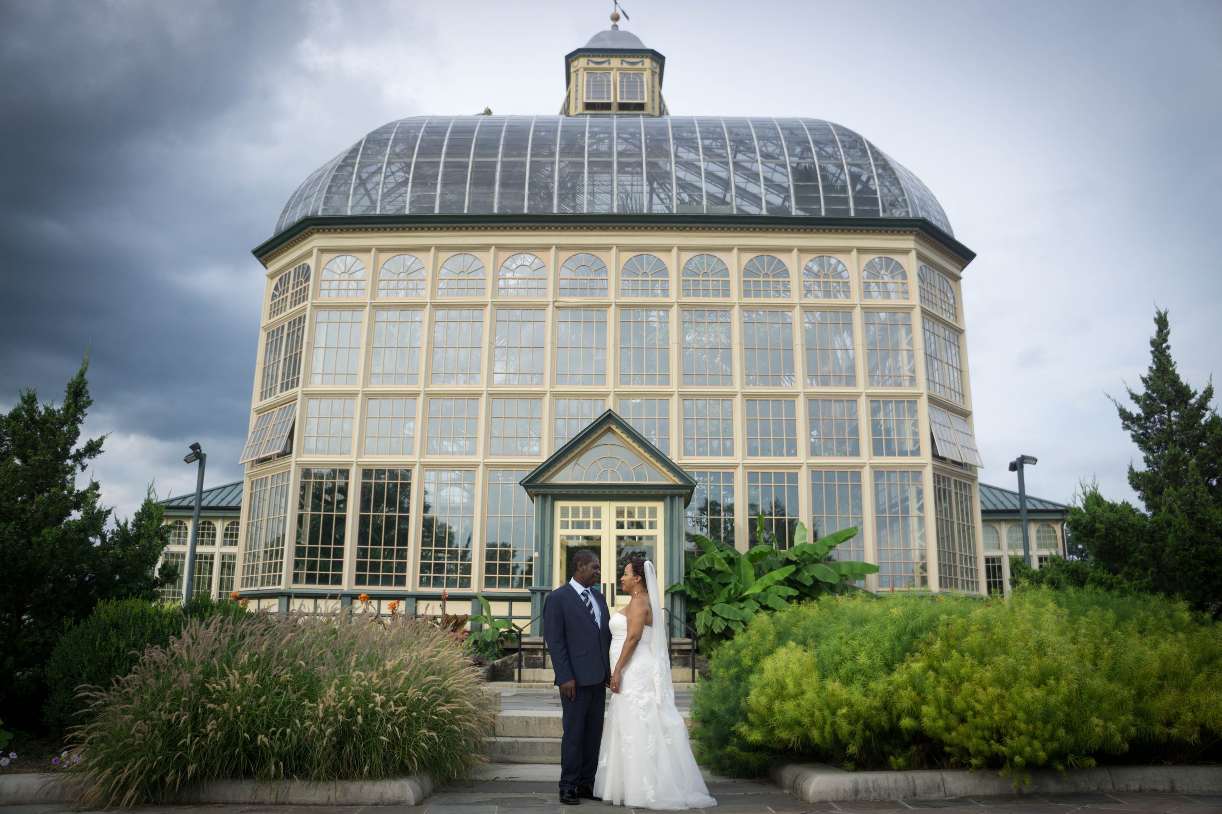 A bride and groom holding hands in front of a large ornate greenhouse, with dark stormy clouds overhead.