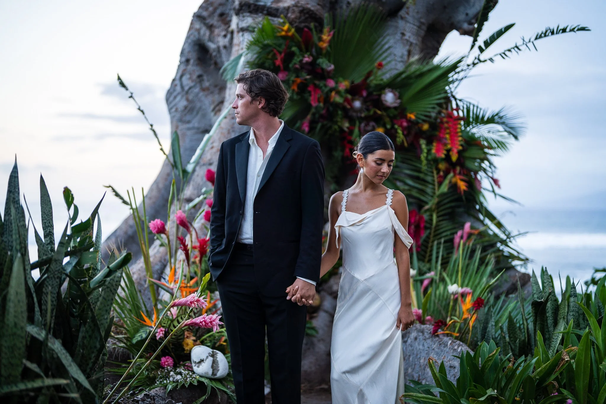 A bride and groom holding hands in front of a floral arch on a beach, with the groom looking to the left and the bride looking down.