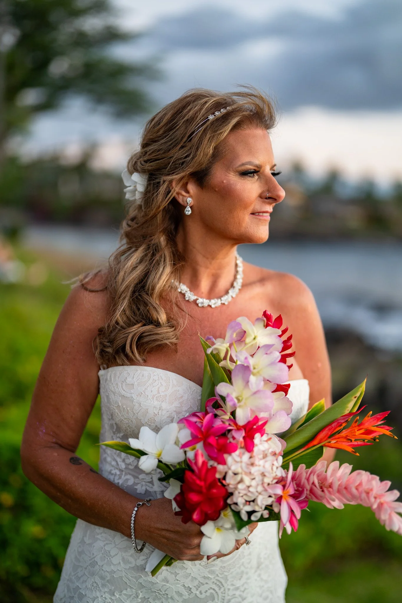 A woman in a white strapless wedding dress holding a bouquet of pink, white, and orange flowers near a body of water with greenery in the background. She has long, wavy hair, jewelry, and is looking to the side.