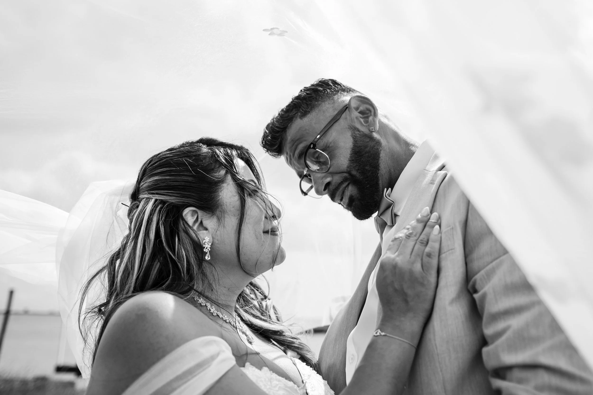 Black and white photograph of a couple sharing an intimate moment, looking into each other's eyes, possibly on their wedding day, outdoors against a cloudy sky.