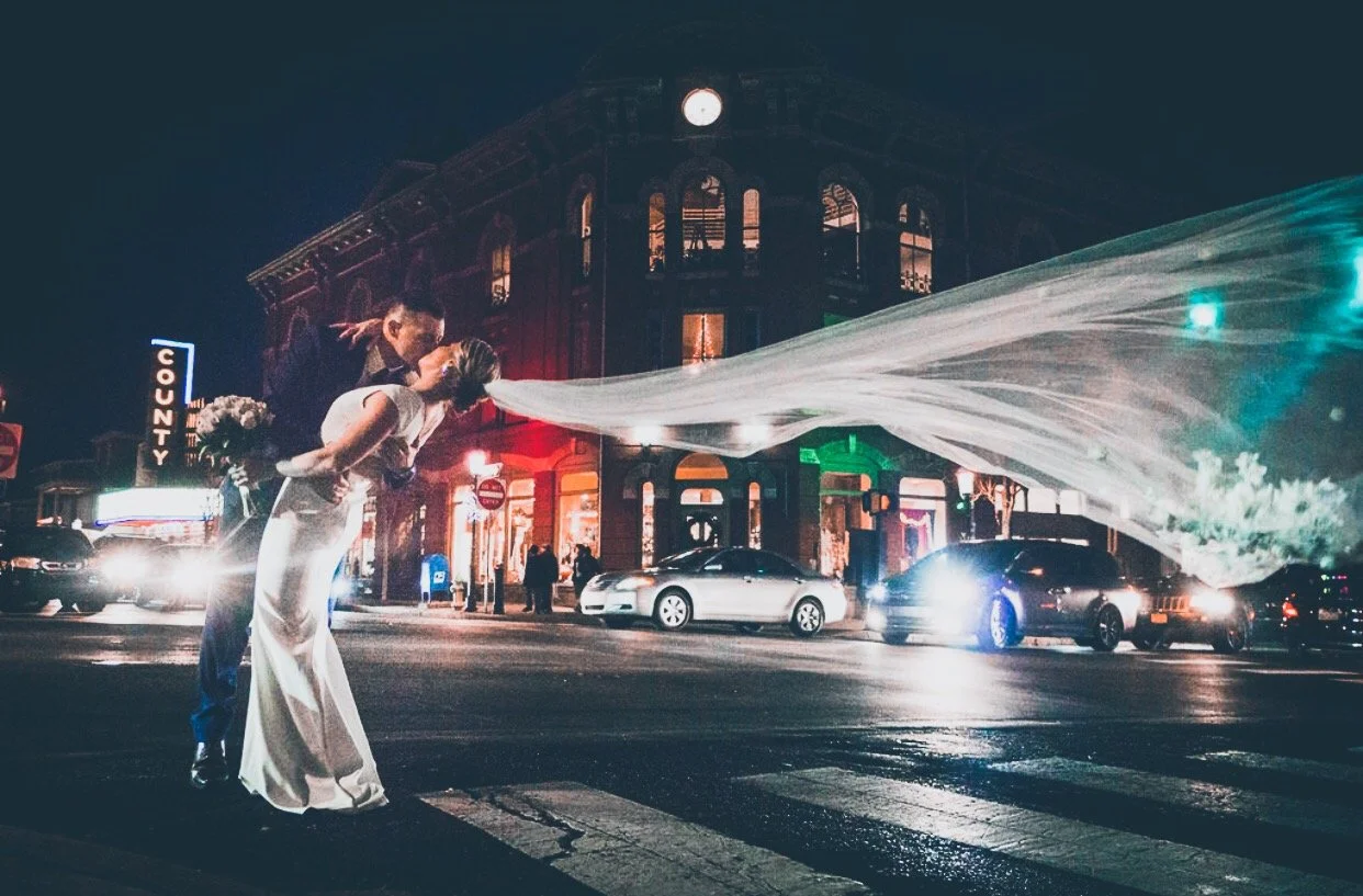 A bride and groom sharing a kiss on a city street at night, with the groom leaning over the bride who is holding a bouquet, and a building with colorful lights in the background.