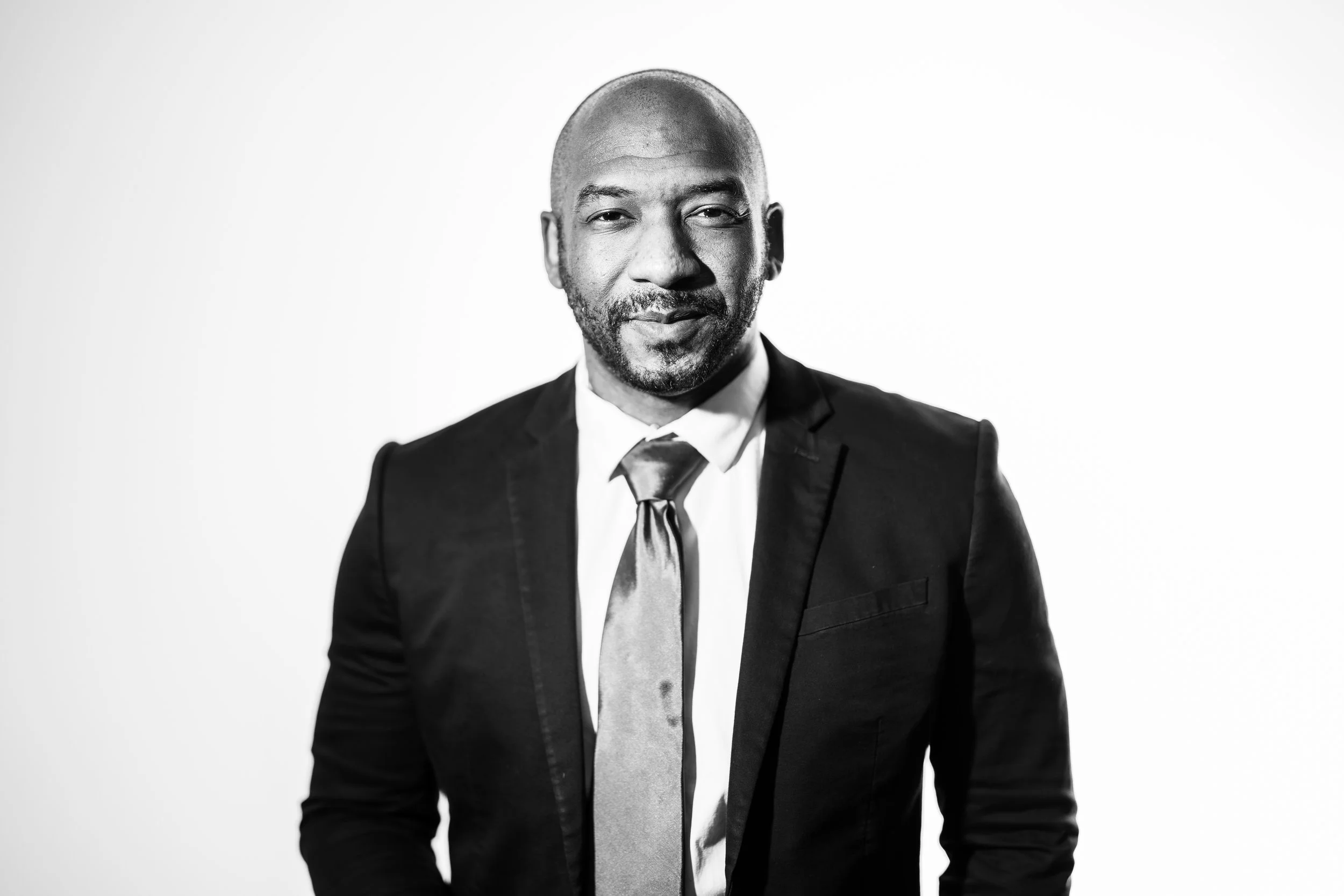 Black and white portrait of a bald man with a beard, wearing a suit jacket, white shirt, and a tie, standing against a plain background.