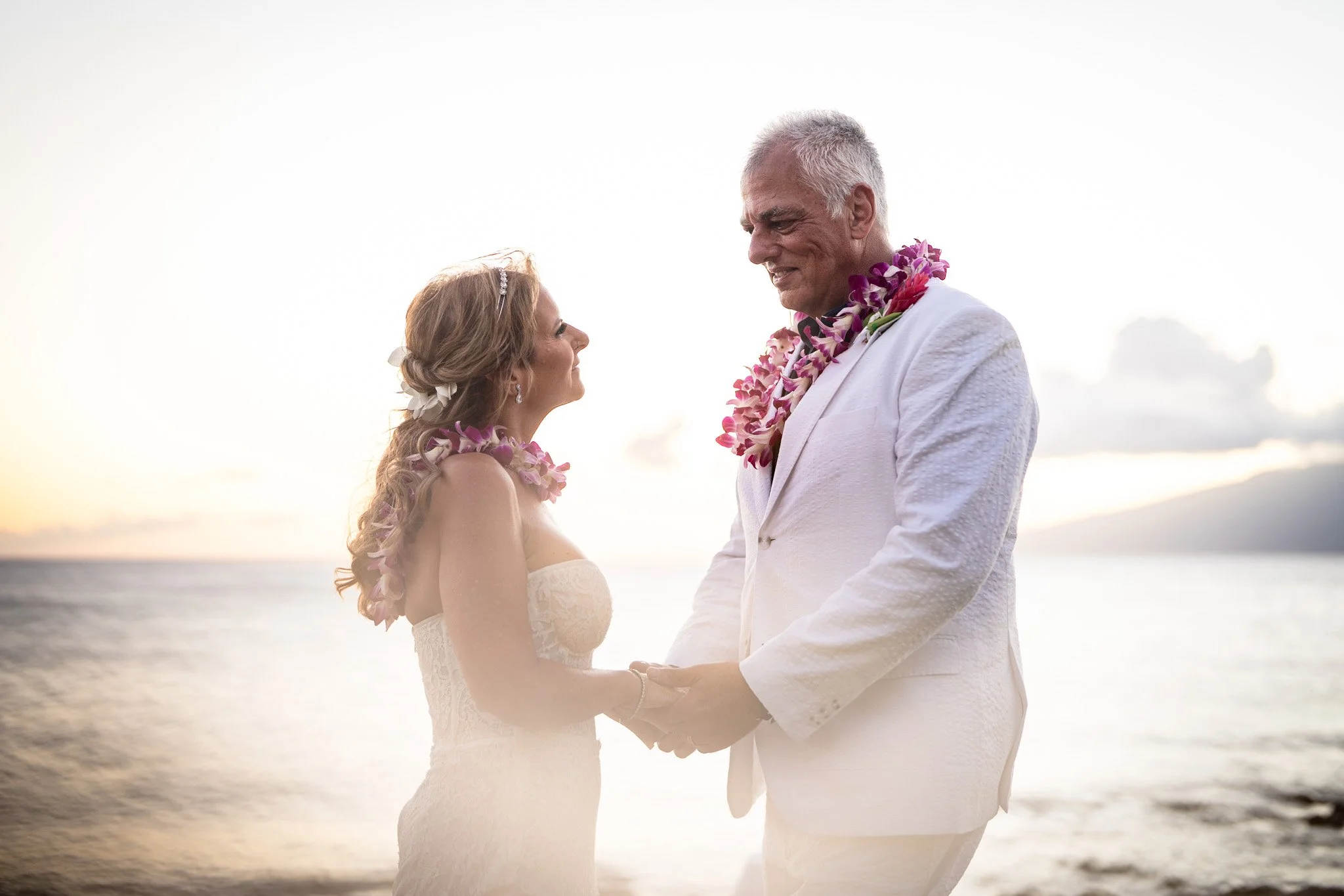 A couple in wedding attire holding hands and gazing at each other on a beach at sunset, wearing leis.