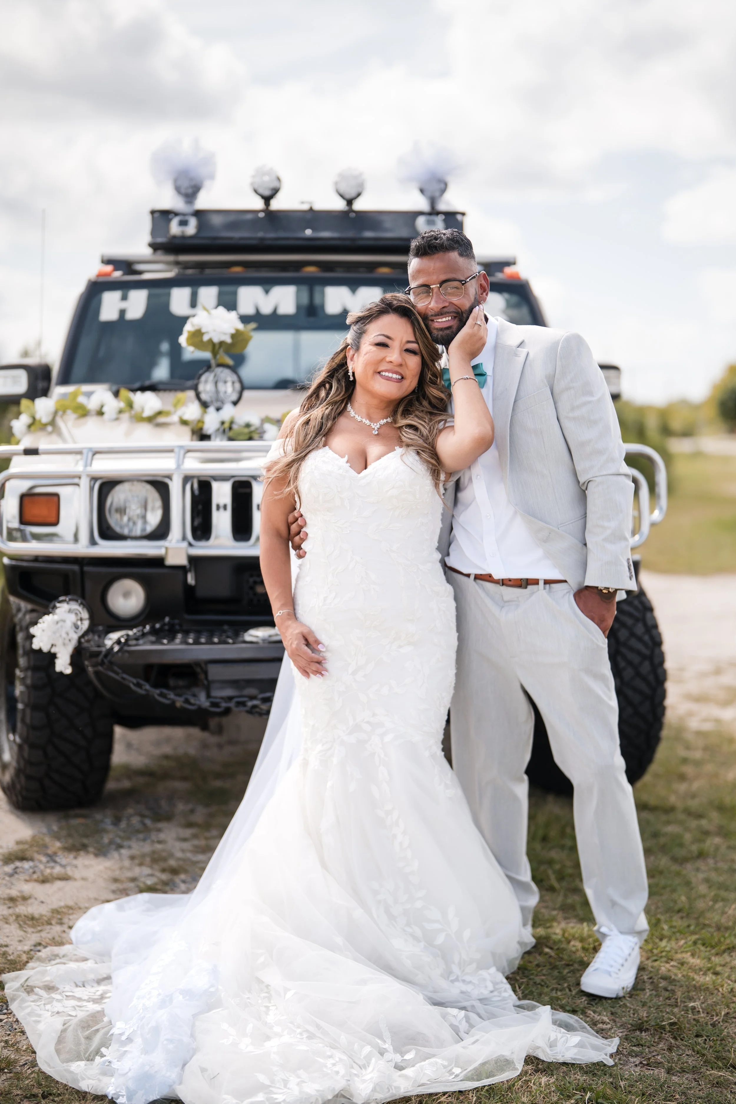 A newlywed couple in wedding attire standing in front of a decorated off-road vehicle, smiling and posing closely.