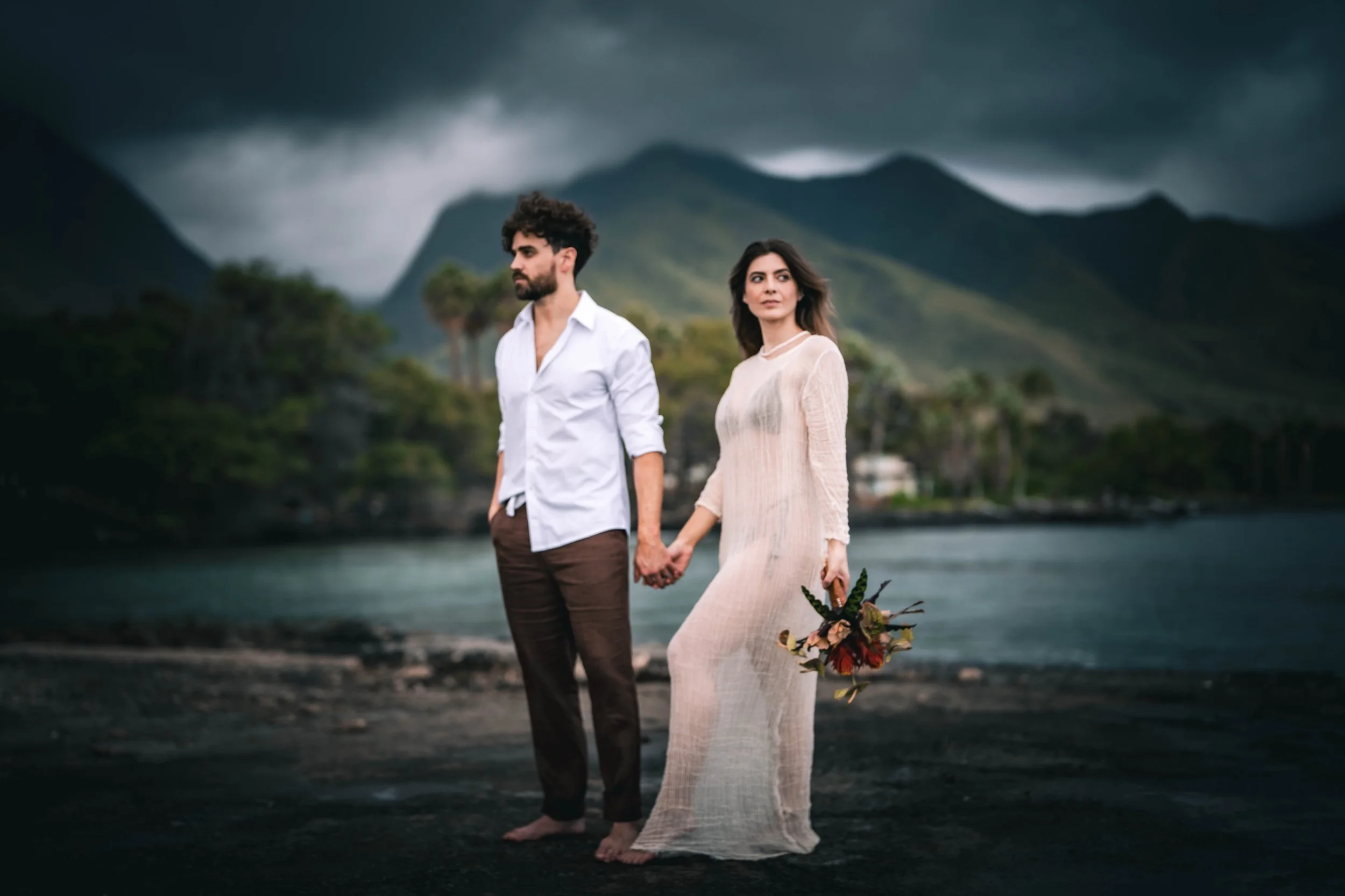 A man and woman holding hands on a dark beach with mountains and stormy sky in the background. The woman holds a bouquet of flowers.