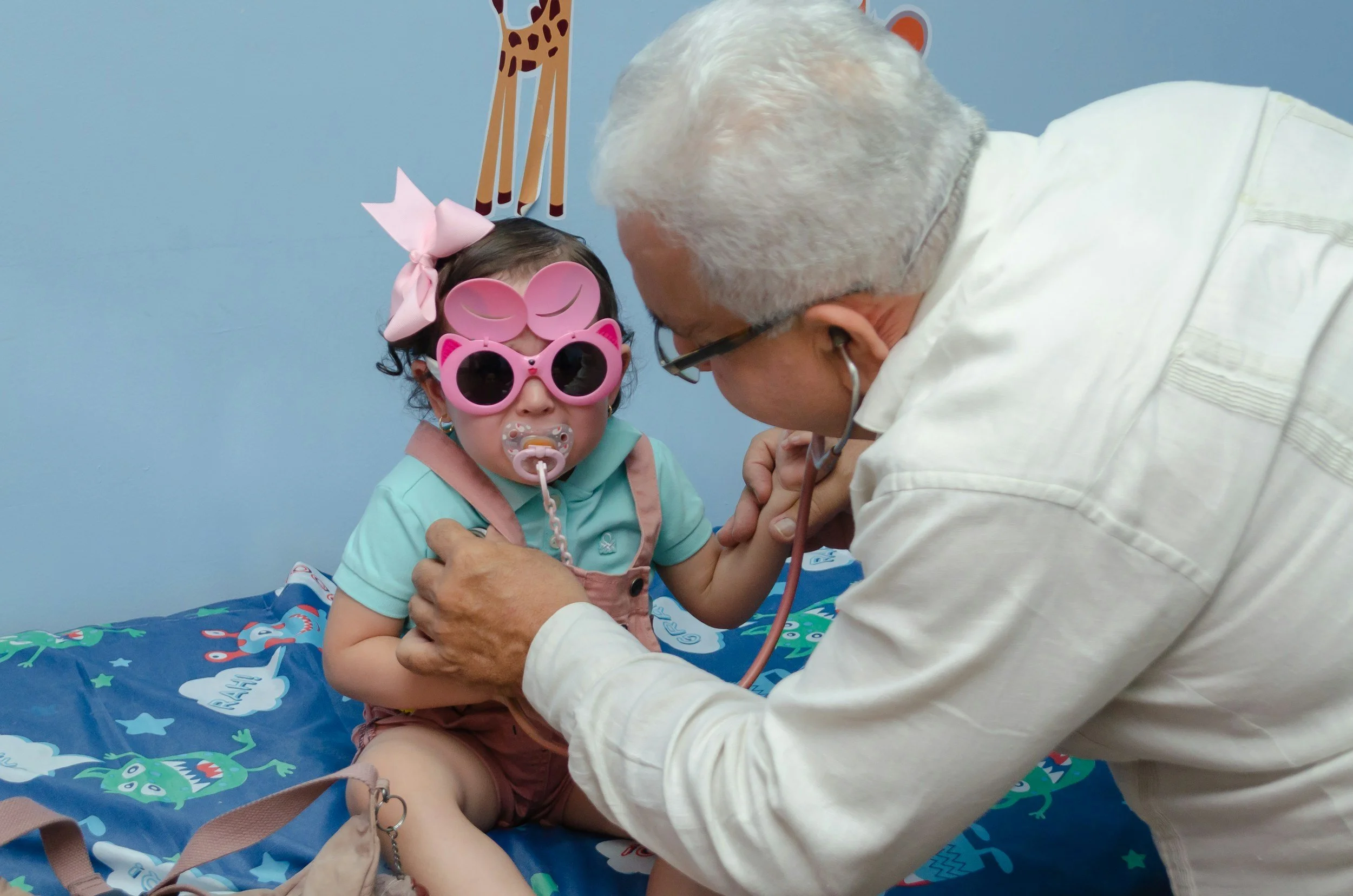 Doctor listening to a child's heartbeat. She's wearing pink glasses and a big pink bow