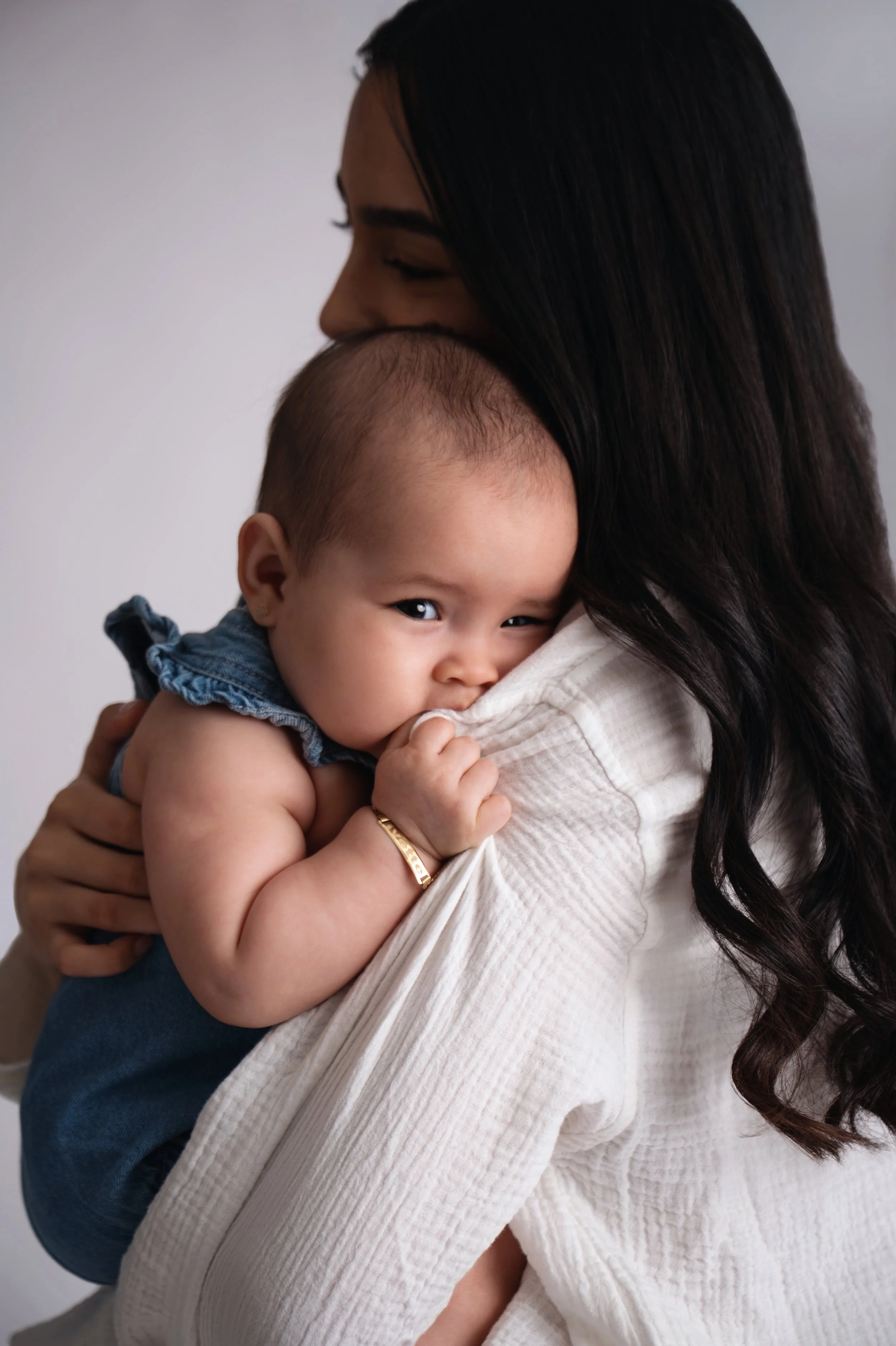 A woman holding a baby close to her shoulder, with the baby's head resting on her chest. The baby is looking at the camera, biting her own hand, wearing a blue outfit, while the woman has long dark hair and is dressed in a white top.