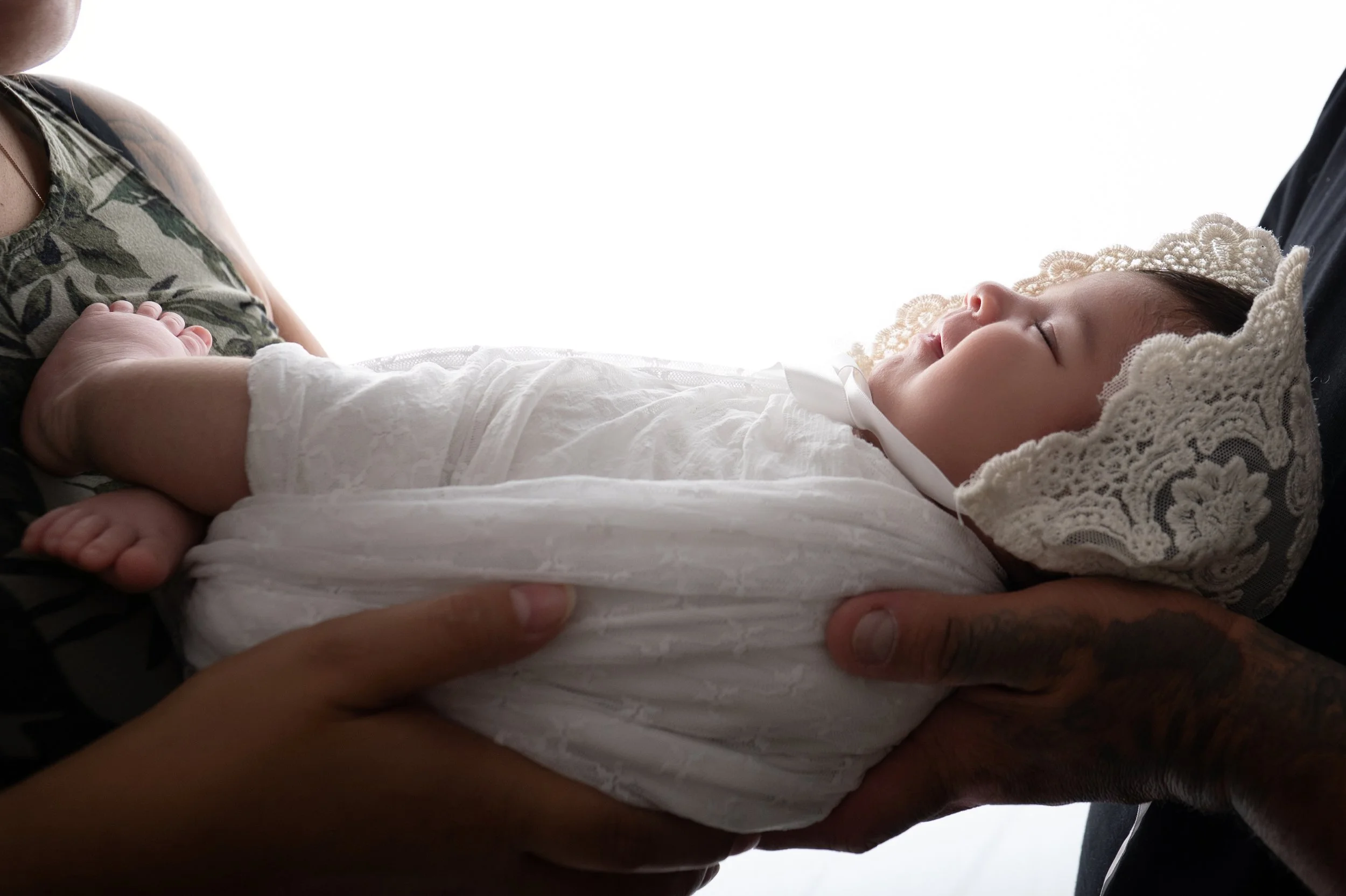 A newborn baby dressed in white, sleeping peacefully with a lace bonnet, being gently held by an adult with tattooed hands.