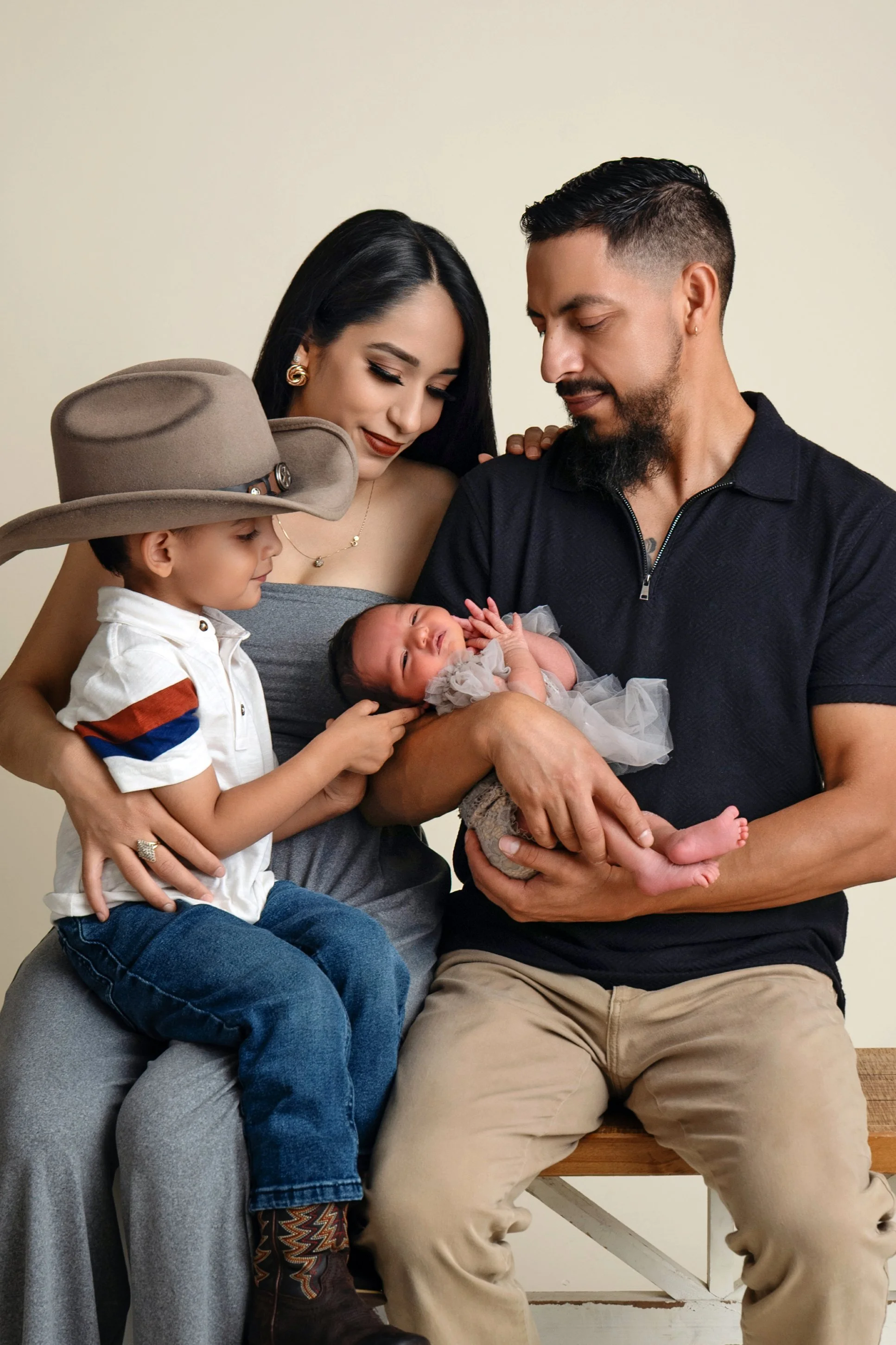 A family of four, including a woman, a man, a young boy, and a newborn baby, gathered together in an indoor setting, with the woman and man holding the baby and the boy touching the baby's hand.