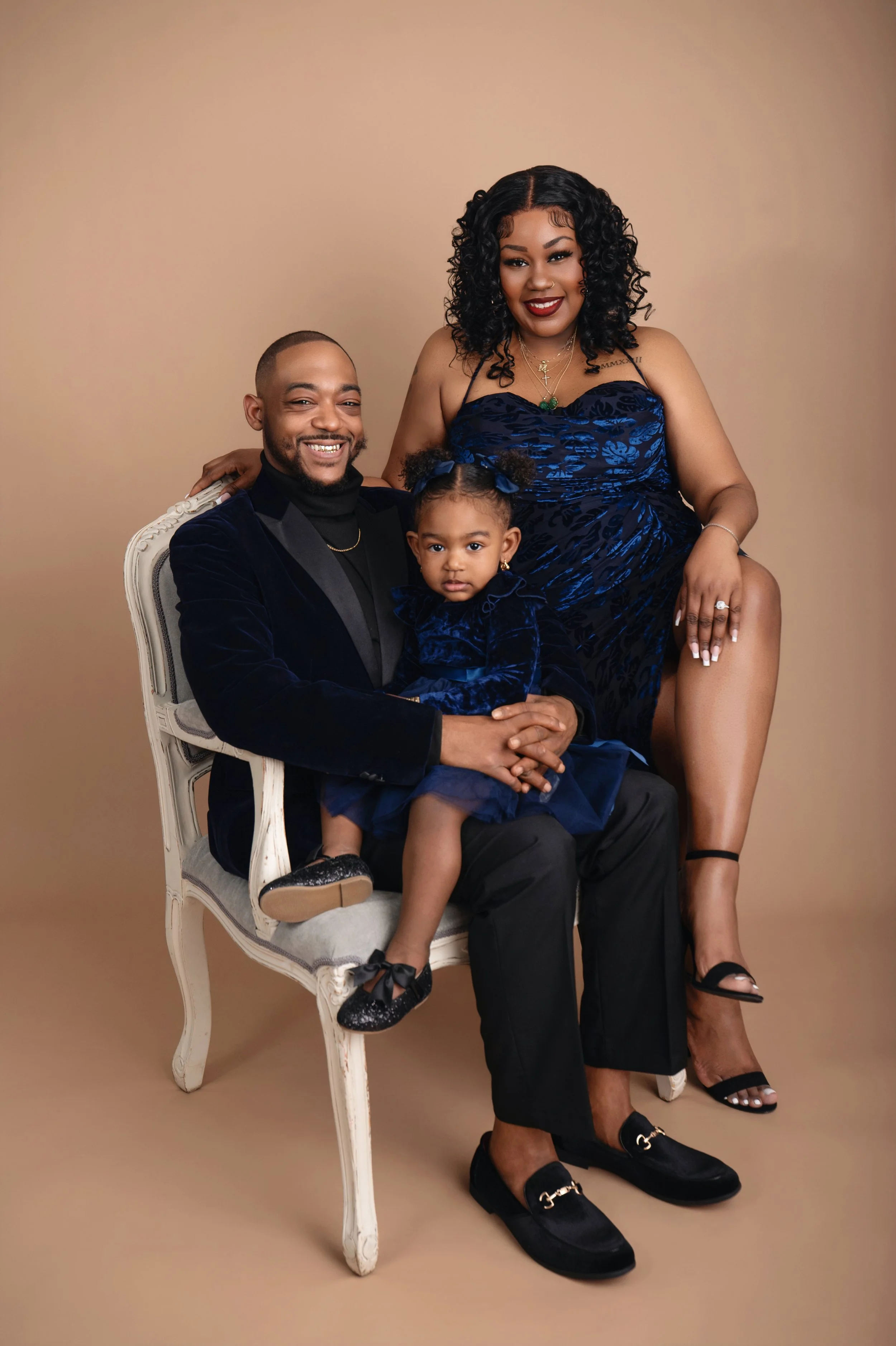 A family of three, including a man, woman, and young girl, dressed in elegant outfits, seated and standing against a beige background for a formal portrait.