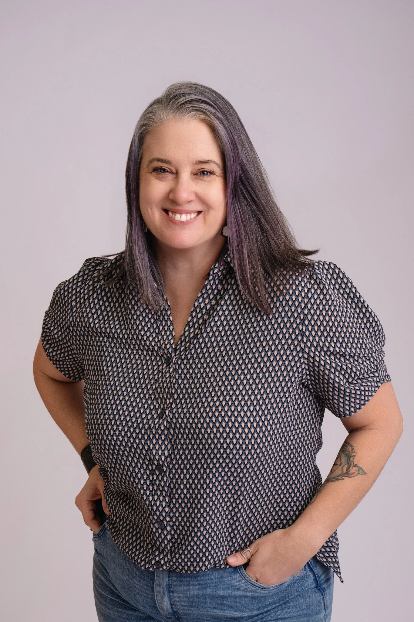 A smiling woman with wavy purple hair, wearing a patterned short-sleeve shirt and blue jeans, standing against a plain light background.