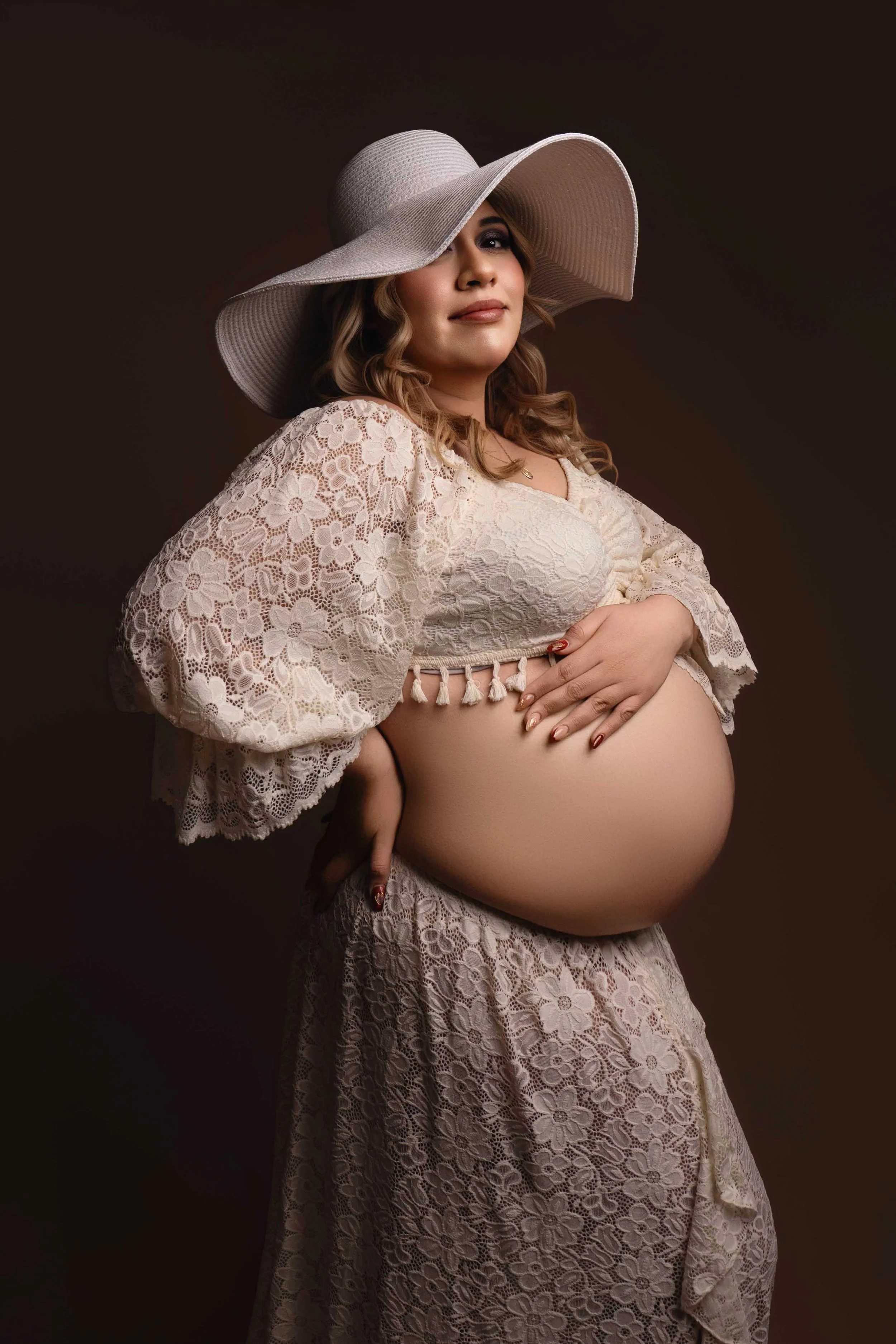 A pregnant woman wearing a large white sunhat and a lace dress with puffed sleeves, gently holding her belly with one hand, posing against a dark background.