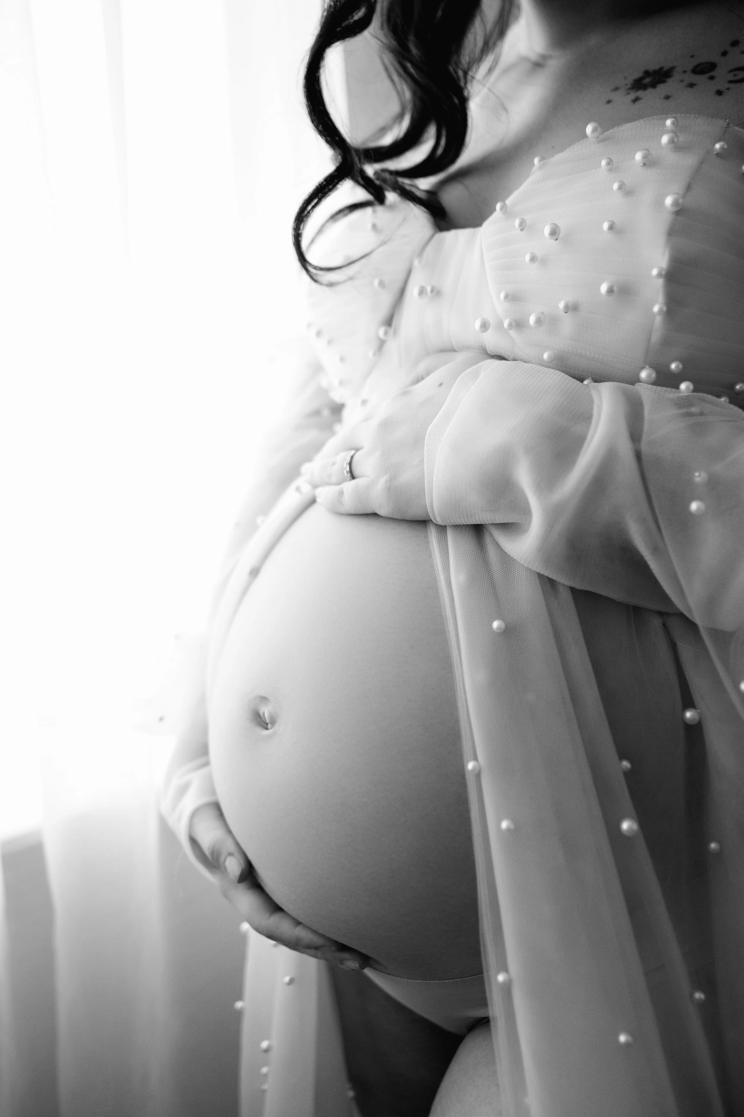 Close-up black and white photo of a pregnant woman holding her belly and wearing a sheer top decorated with pearls.
