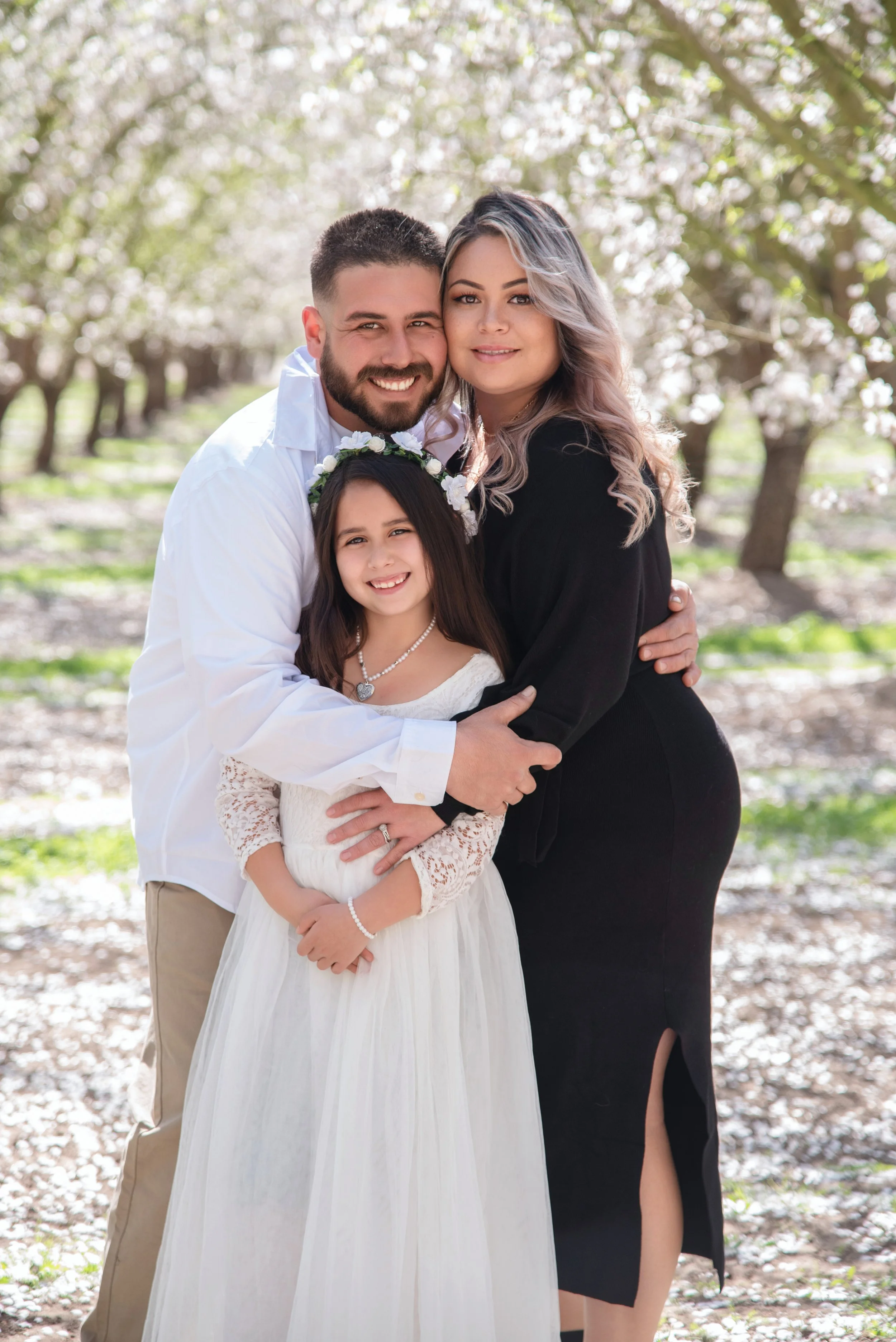 Family of 3 lovingly hugging together in the middle of a almond blossom orchard locaded in Modesto CA
