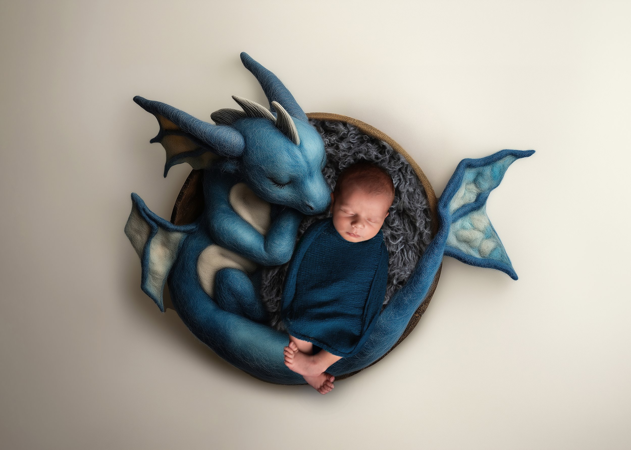 A newborn baby wrapped in a dark blue blanket, sleeping on a fluffy gray surface inside a round basket, surrounded by a blue dragon plush toy.