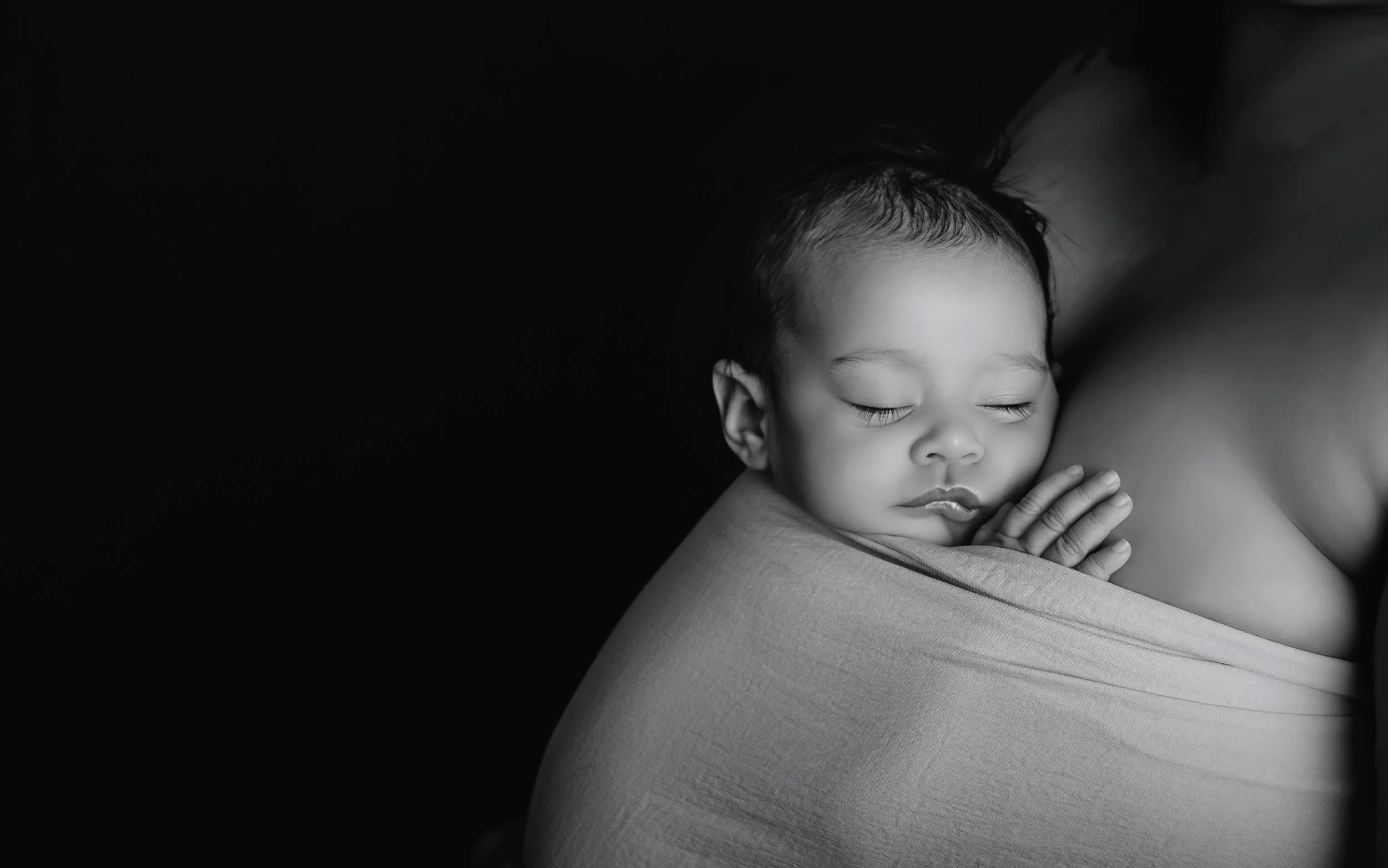 Black and white photo of a sleeping baby cuddled against a person's chest, wrapped in a blanket.