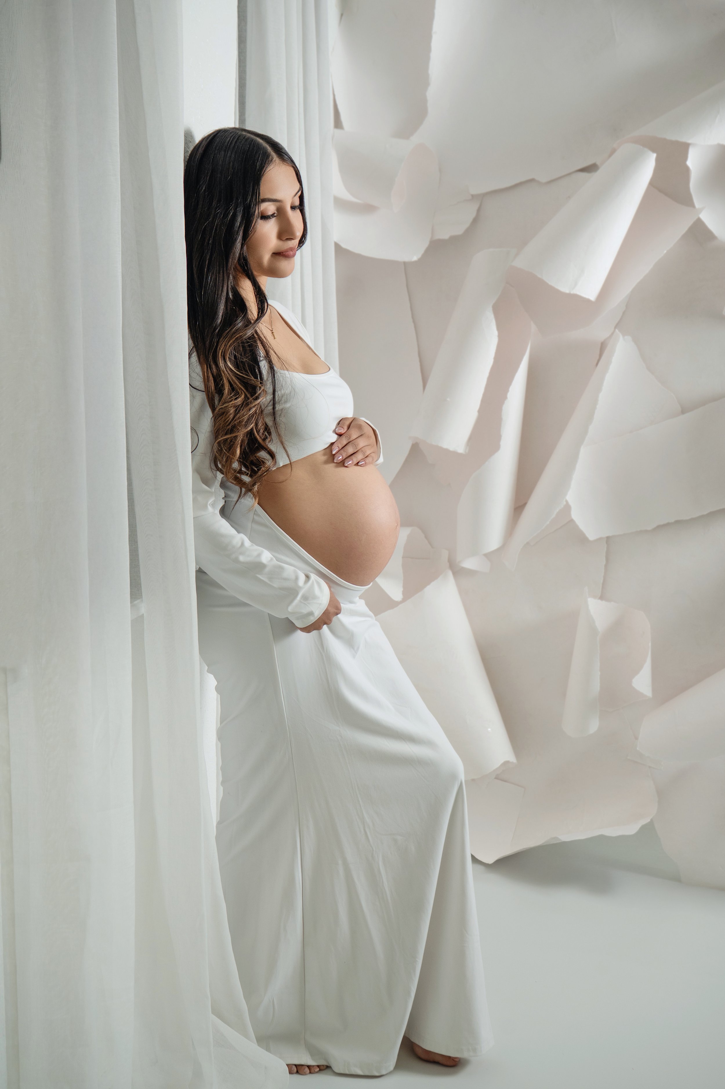 Pregnant woman in white outfit standing by sheer curtains with abstract white paper background.