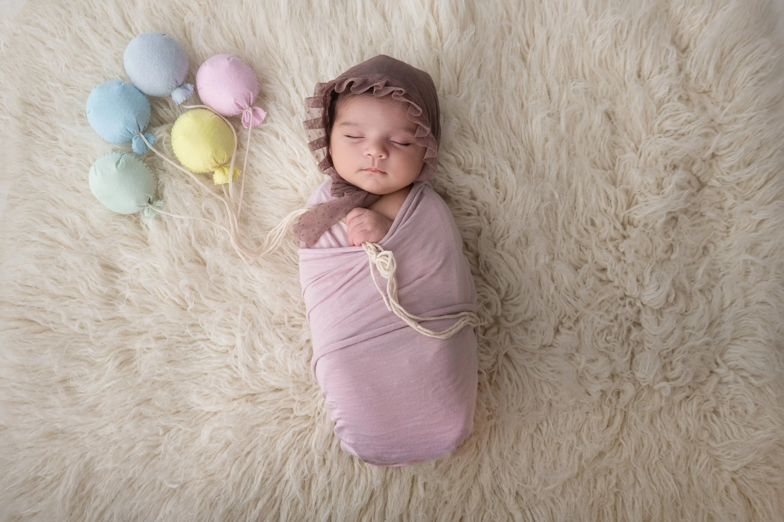 Newborn baby wrapped in soft pink fabric, posed on a white fur blanket and holding pastel balloons in a professional studio newborn photography session in Modesto, California.