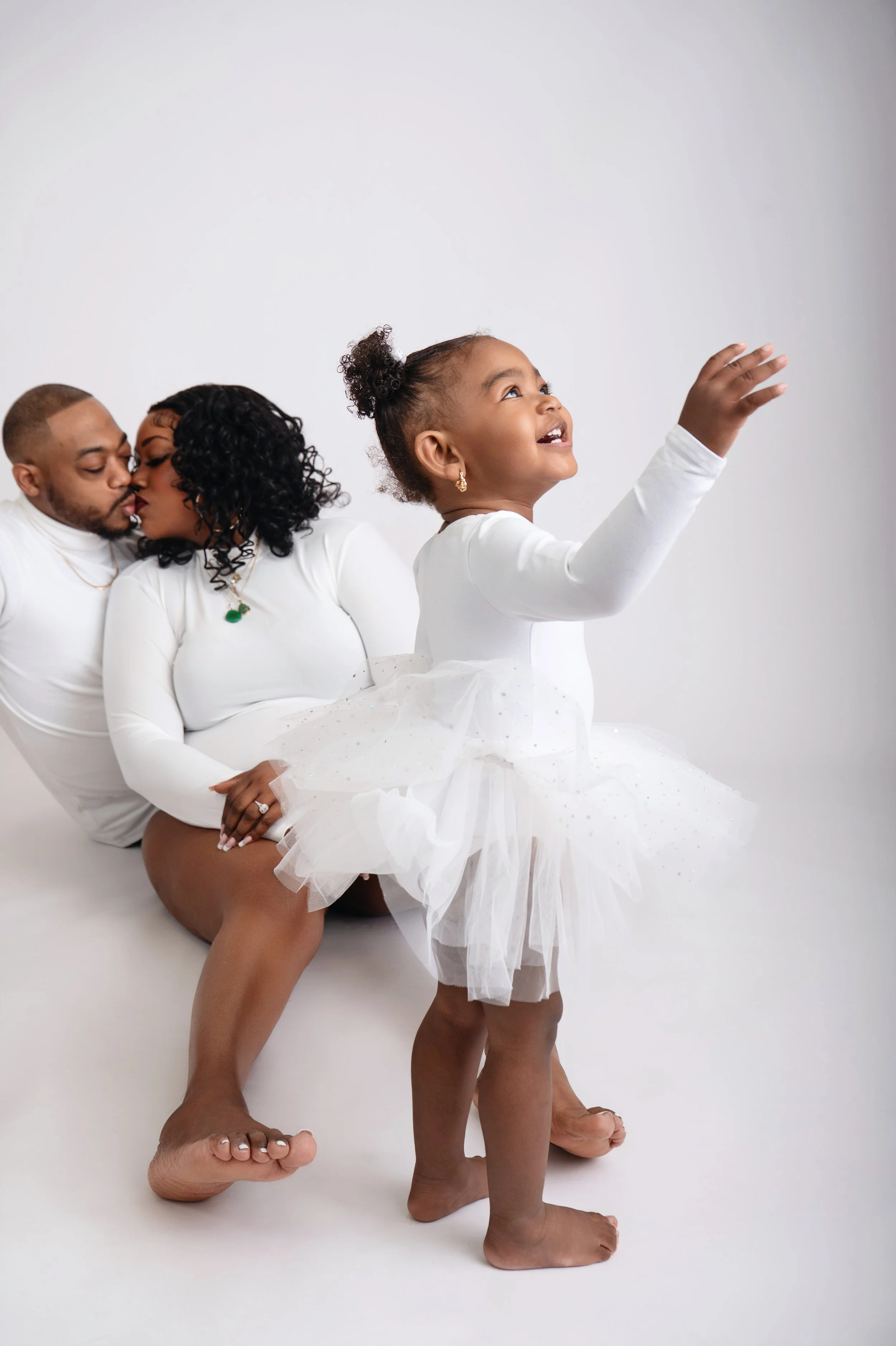 A happy little girl in a white dress reaching upward, with two adults in white clothing sitting behind her on a white background, sharing a moment.