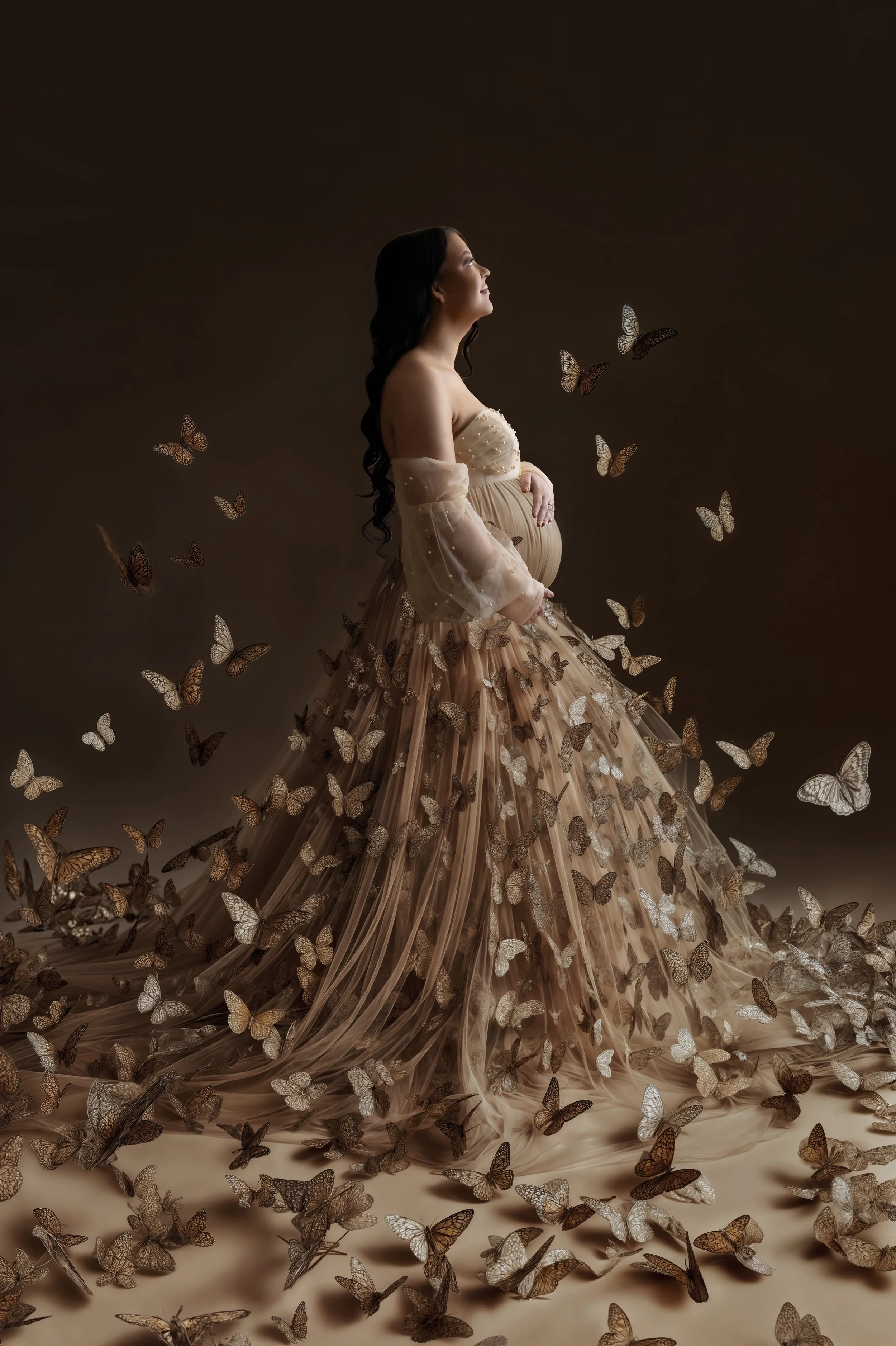 Pregnant mother softly lit from the side, gazing up while surrounded by delicate butterflies during a fine art maternity photography session.