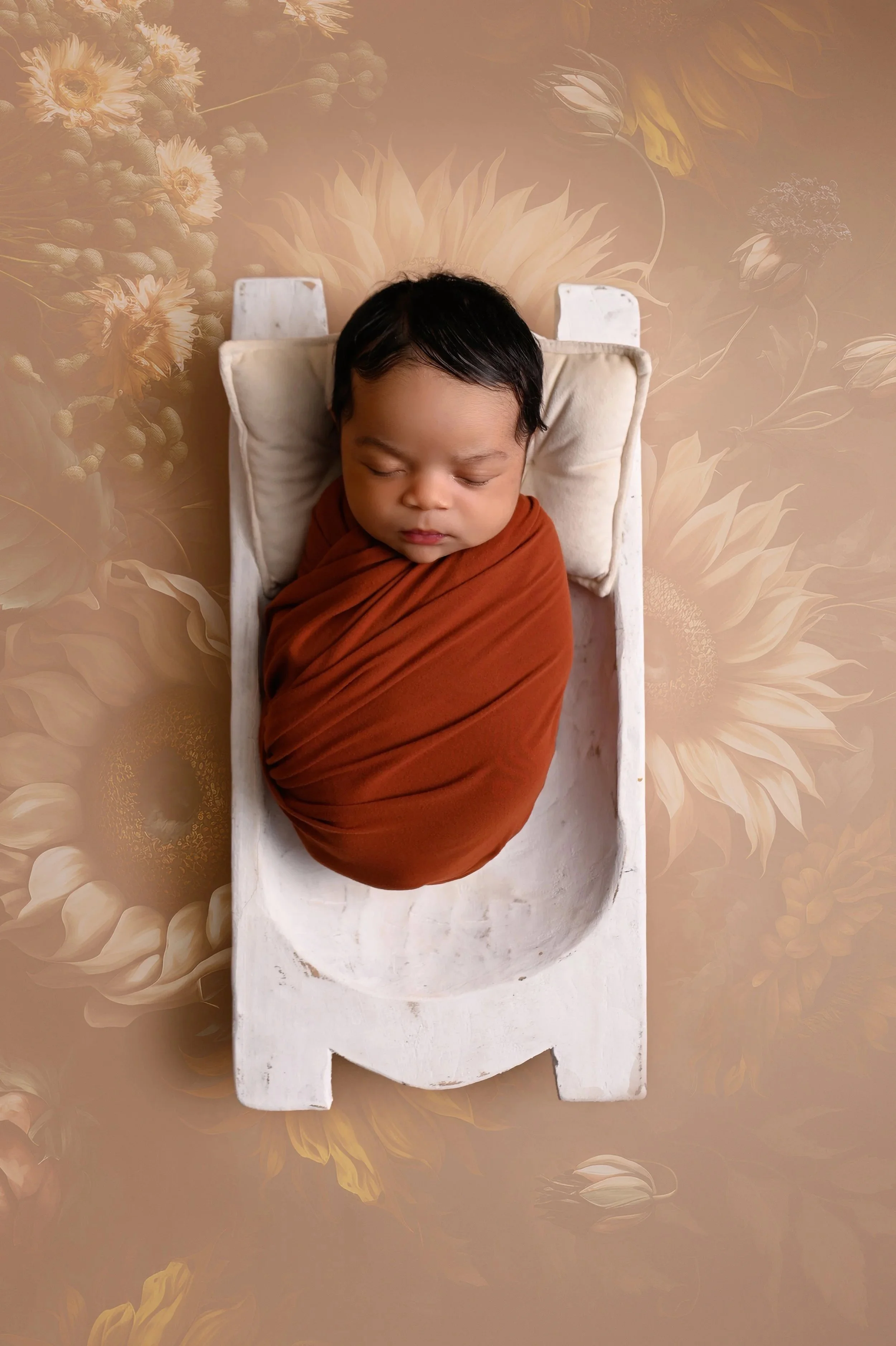 A baby wrapped in an orange swaddle sleeping in a white wooden cradle on a floral background.