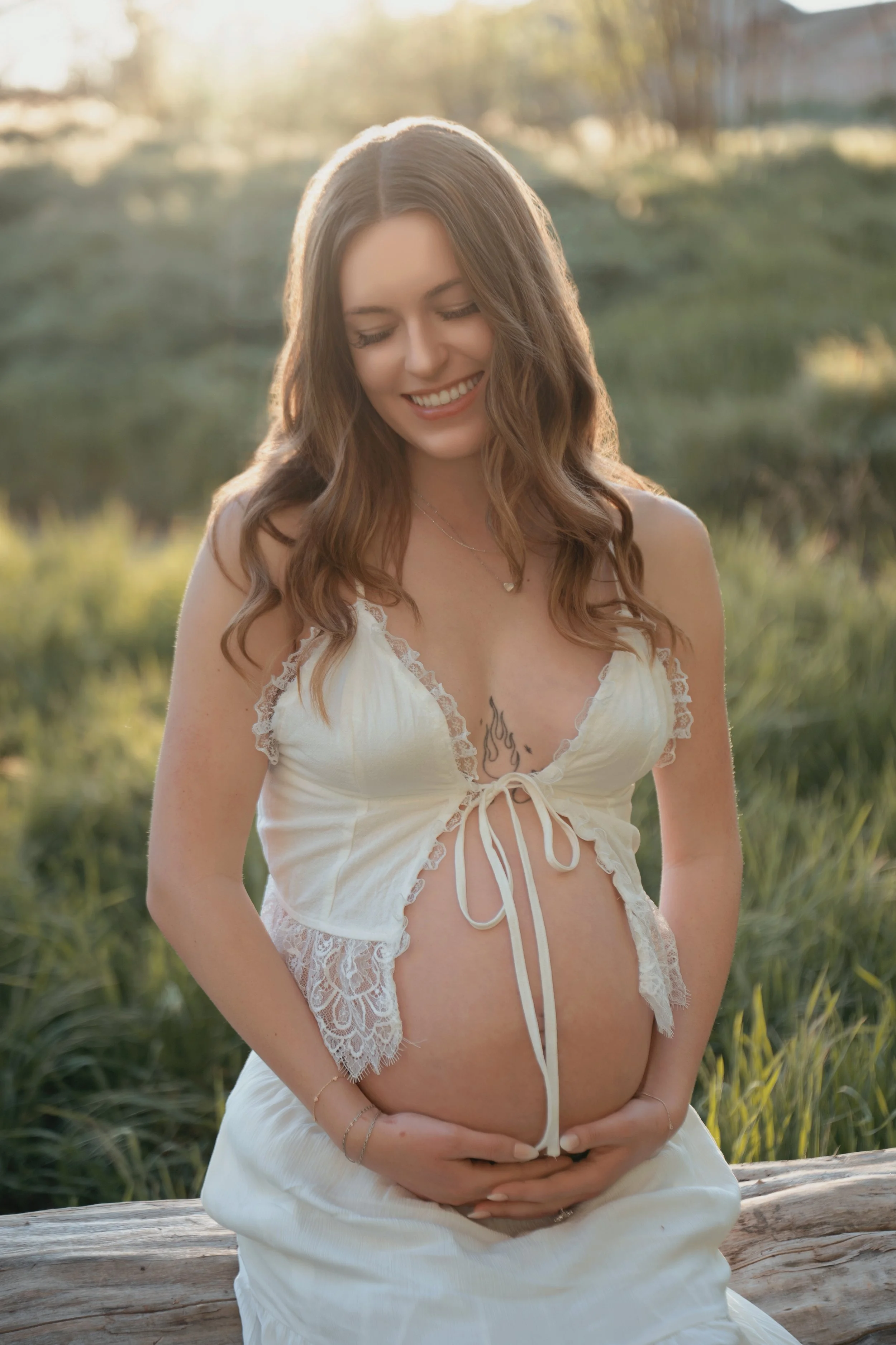 A smiling woman with wavy brown hair sitting on a log outdoors during golden hour, wearing a white lace and satin wrap dress, with a tattoo visible on her chest, surrounded by green blurred background of trees and grass.