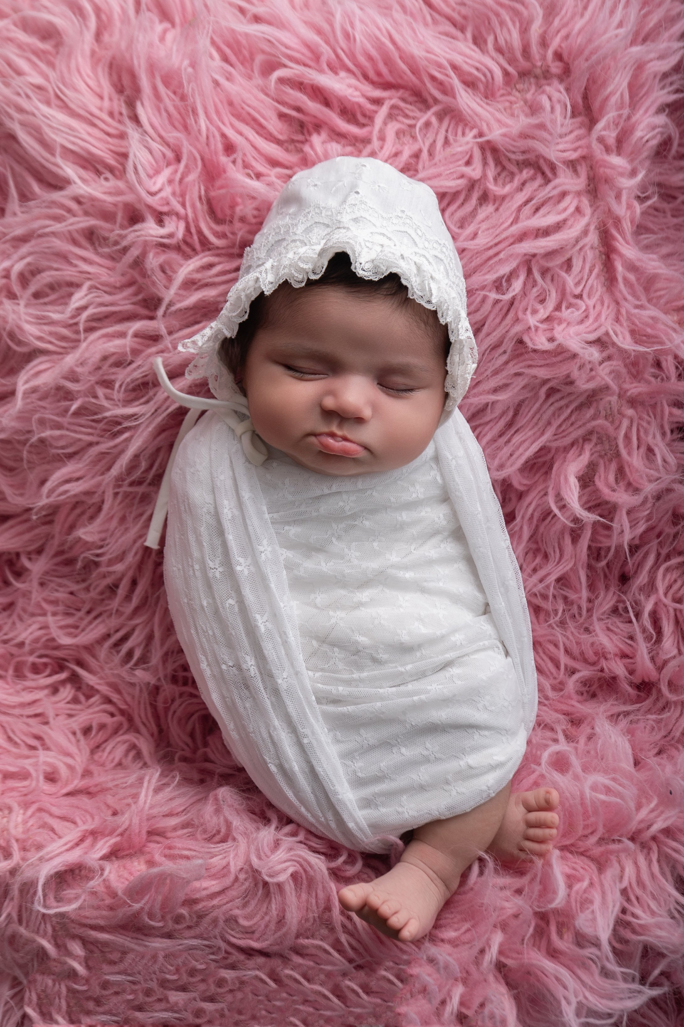 A sleeping baby girl wrapped in a white blanket with a matching bonnet, lying on a pink fluffy blanket.