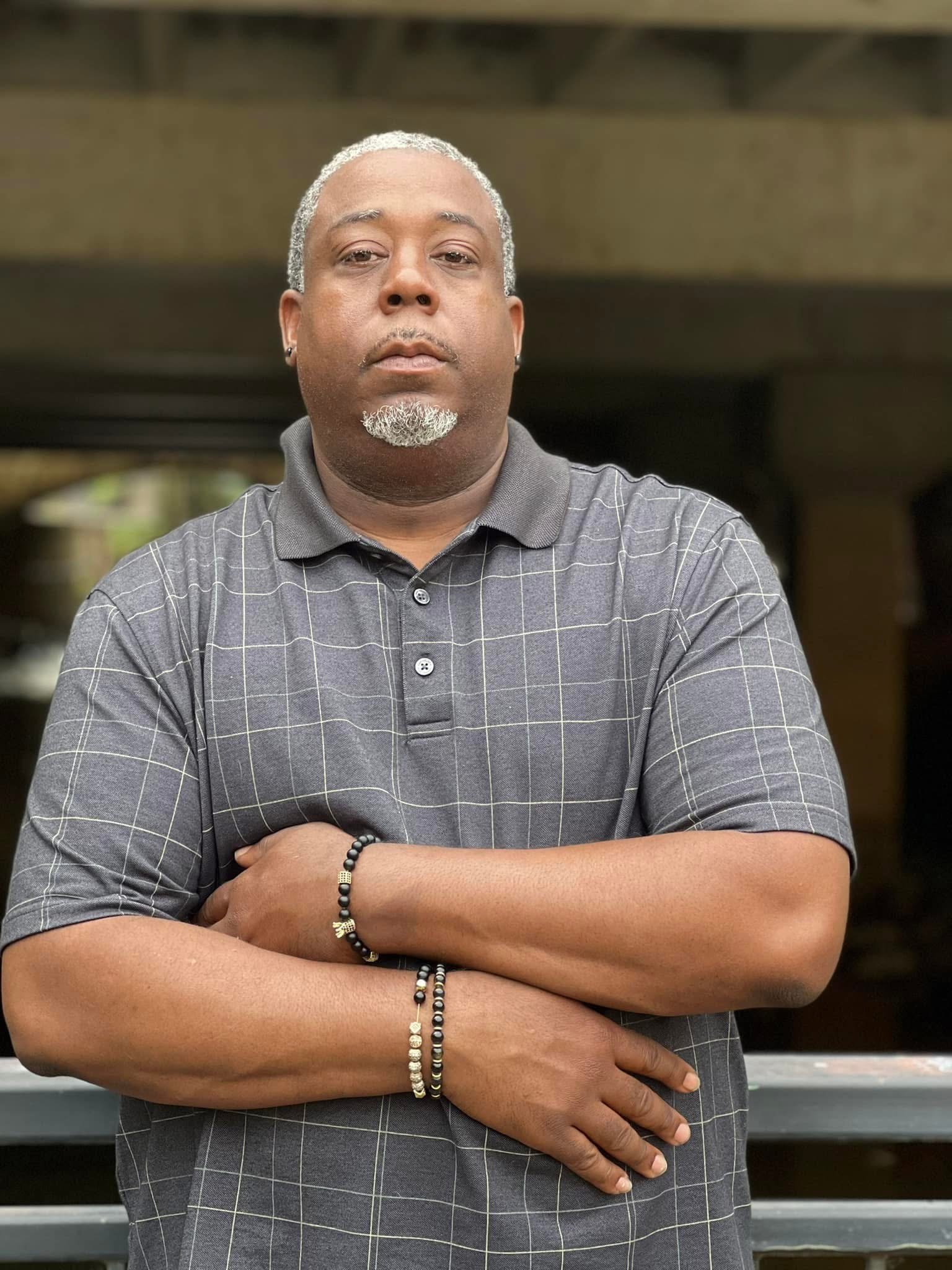 A man with gray hair and a beard standing outdoors with his arms crossed, wearing a gray checkered polo shirt and bracelets.