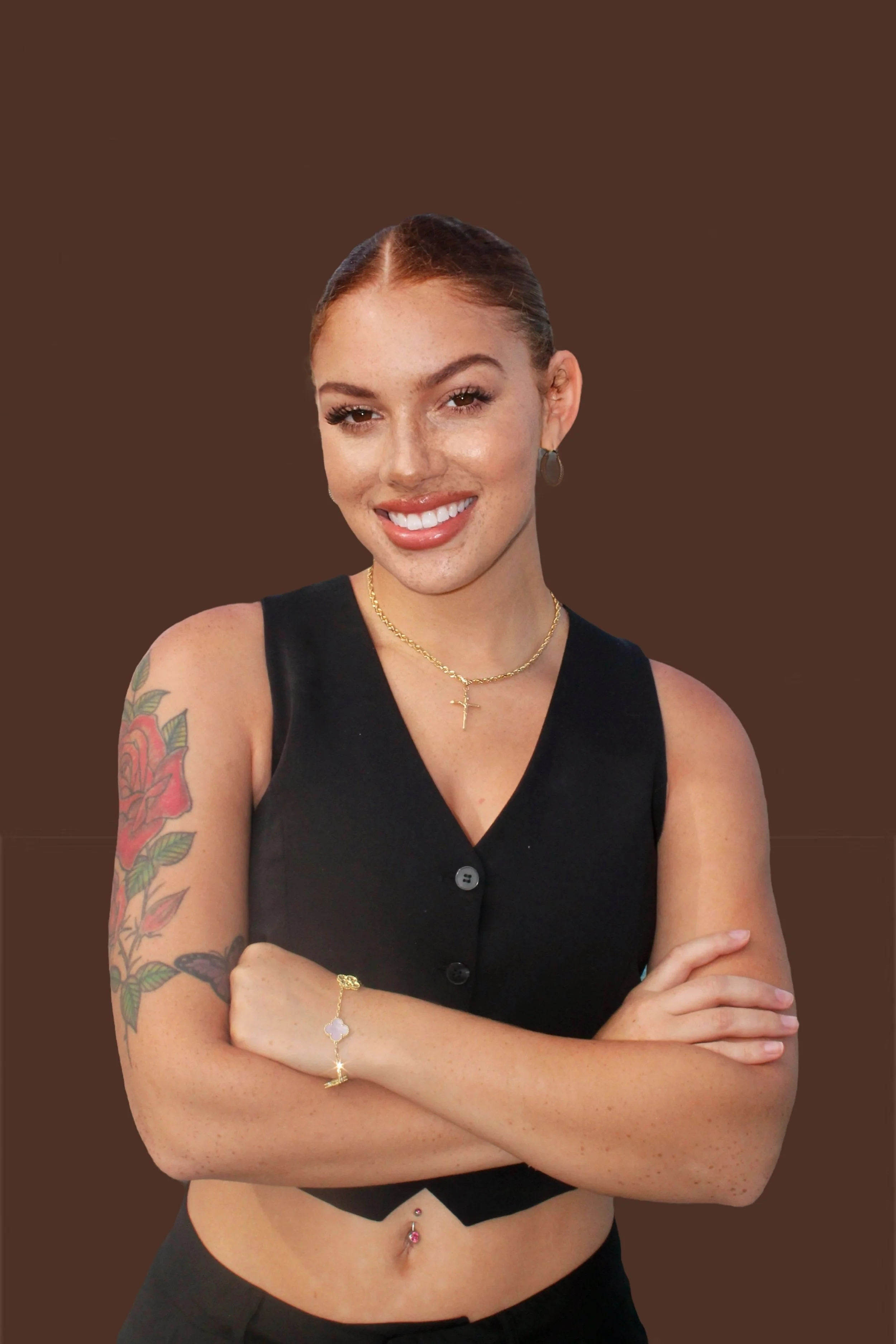 A young woman with red hair, freckles, wearing a black sleeveless top, gold jewelry, and a tattoo on her left arm, smiling with arms crossed against a solid brown background.