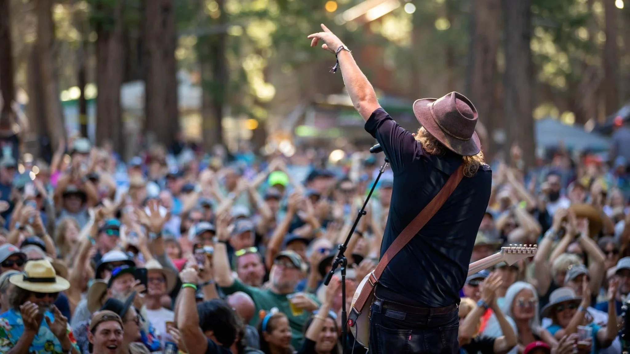 Musician wearing a brown hat and black shirt playing guitar on stage with a large outdoor audience in the background.